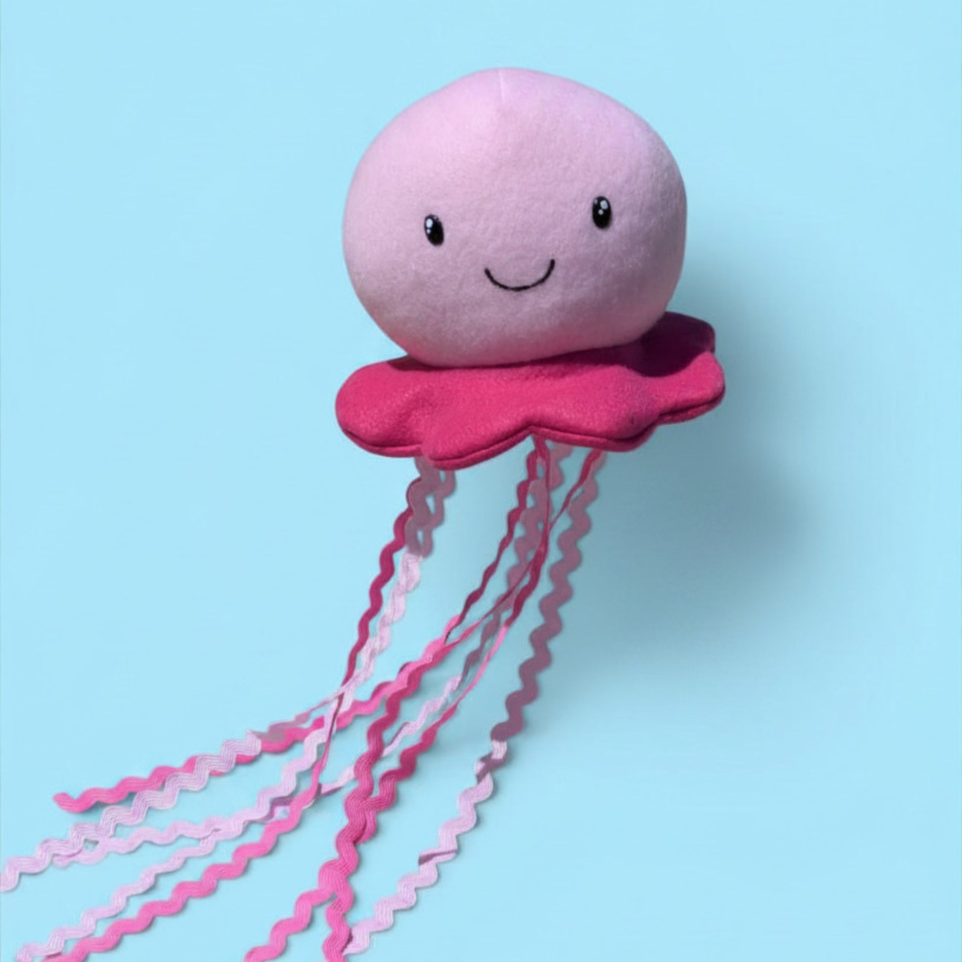 A pink plush jellyfish toy with a smiling face, a darker pink flared bottom, and long wavy pink tentacles, shown against a light blue background.