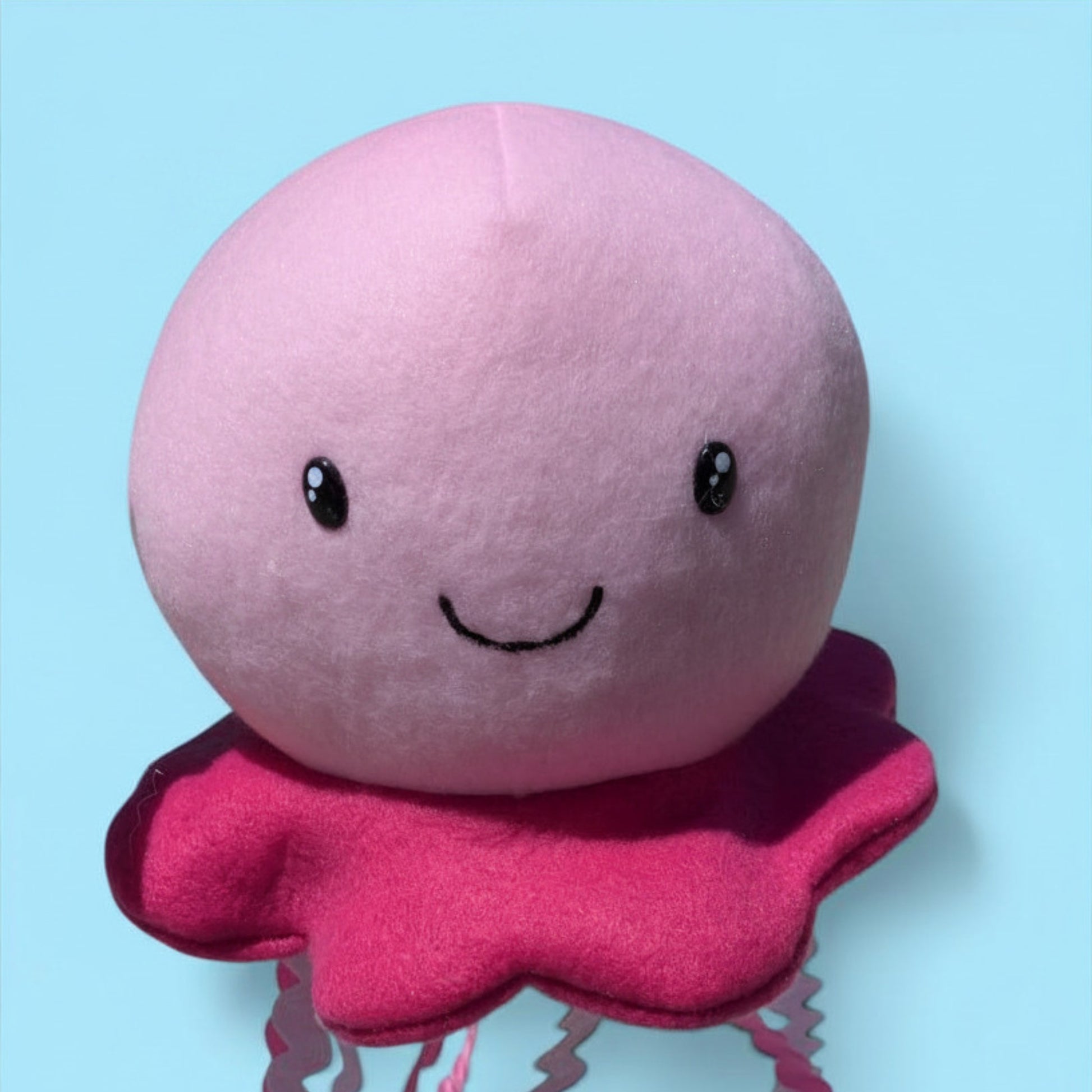 A cute pink jellyfish plush toy with a smiling face and darker pink tentacles, set against a solid light blue background.