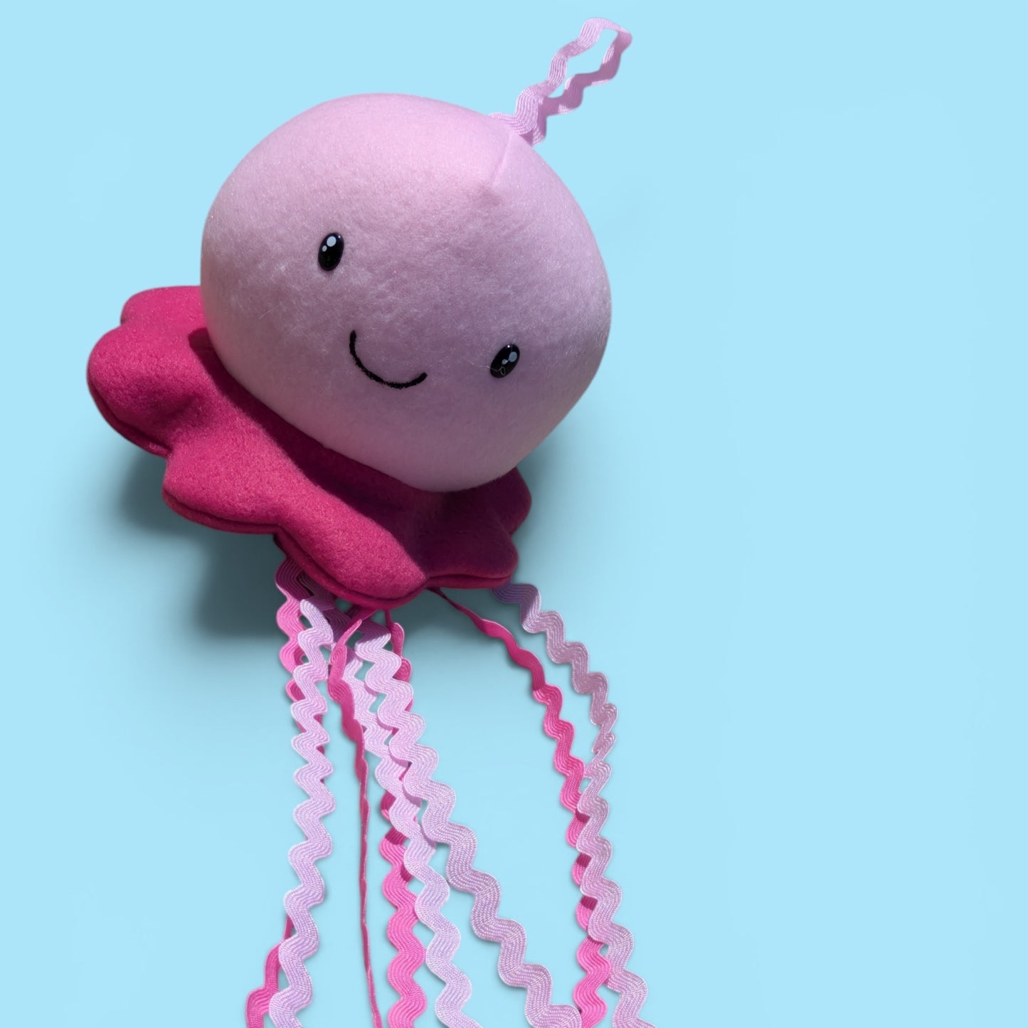 A pink plush jellyfish toy with a smiling face, a darker pink ruffled bottom, and long wavy pink tentacles, photographed against a light blue background.