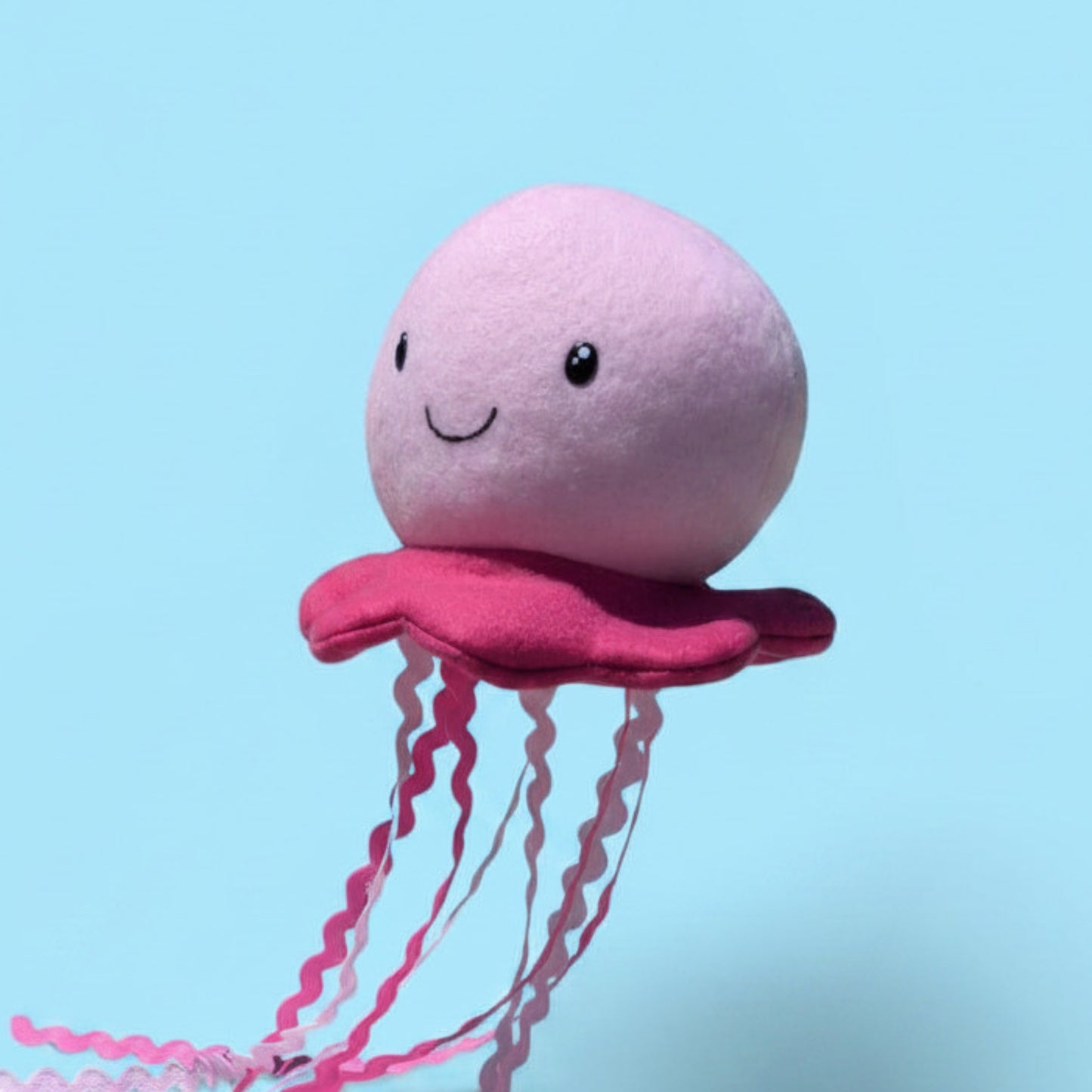 A cute pink jellyfish plush toy with a smiling face and wavy tentacles, set against a solid light blue background.
