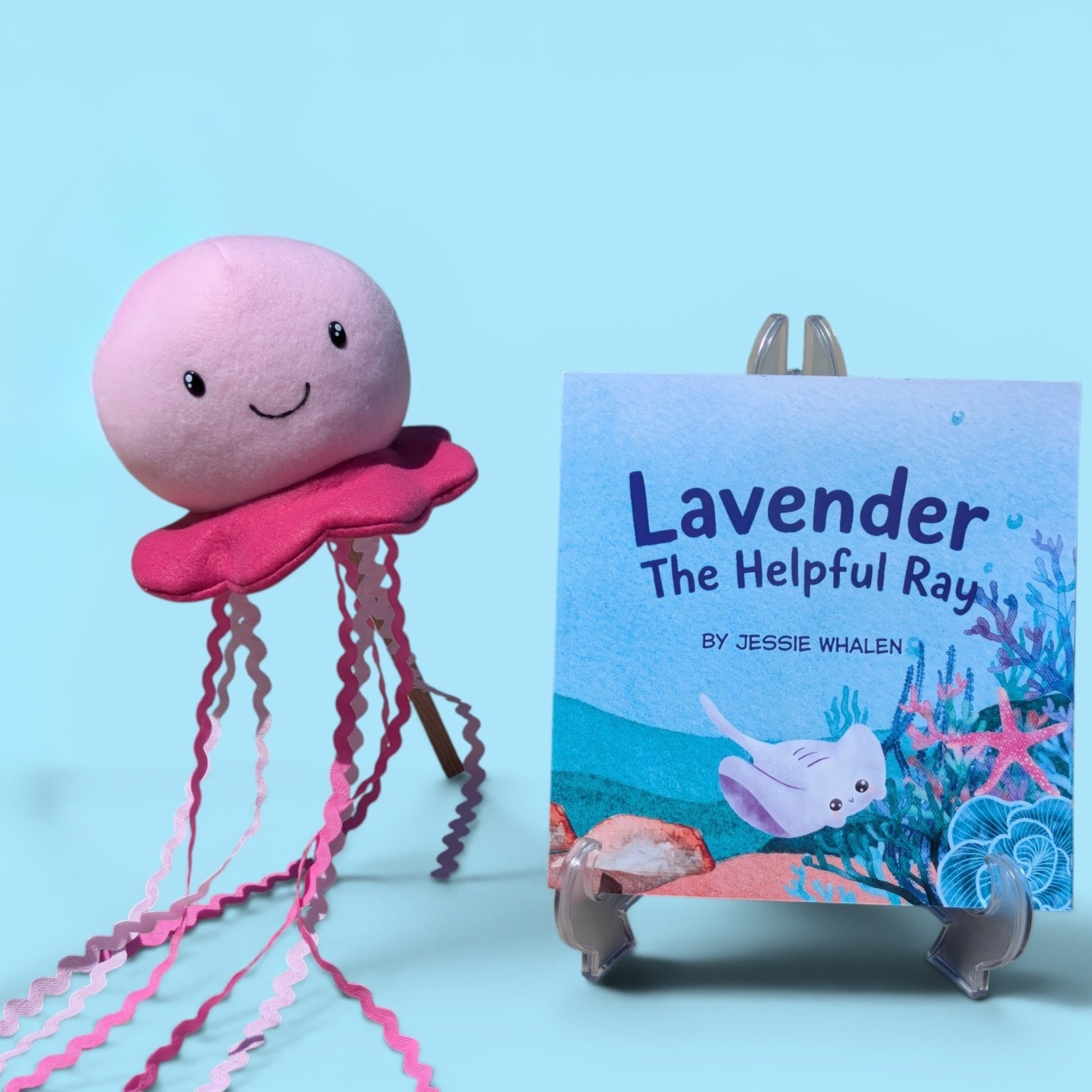A pink plush jellyfish toy with long wavy pink tentacles stands beside a children's book titled "Lavender The Helpful Ray" by Jessie Whalen, displayed on a small stand against a light blue background.