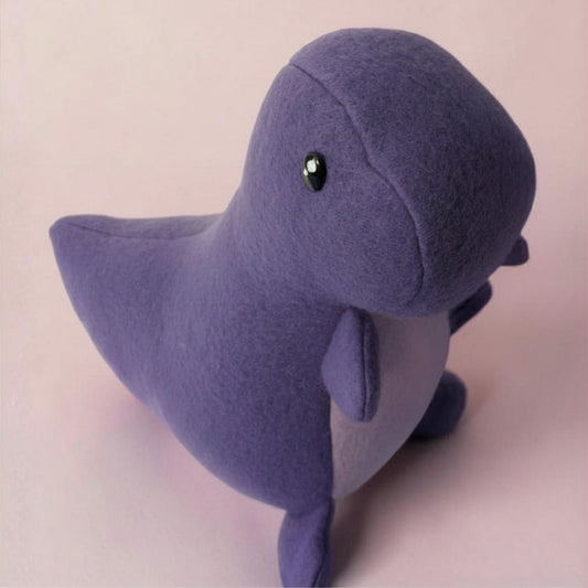 A small plush dinosaur toy made of soft purple fleece fabric, featuring a black safety eye, set against a solid pale pink background.