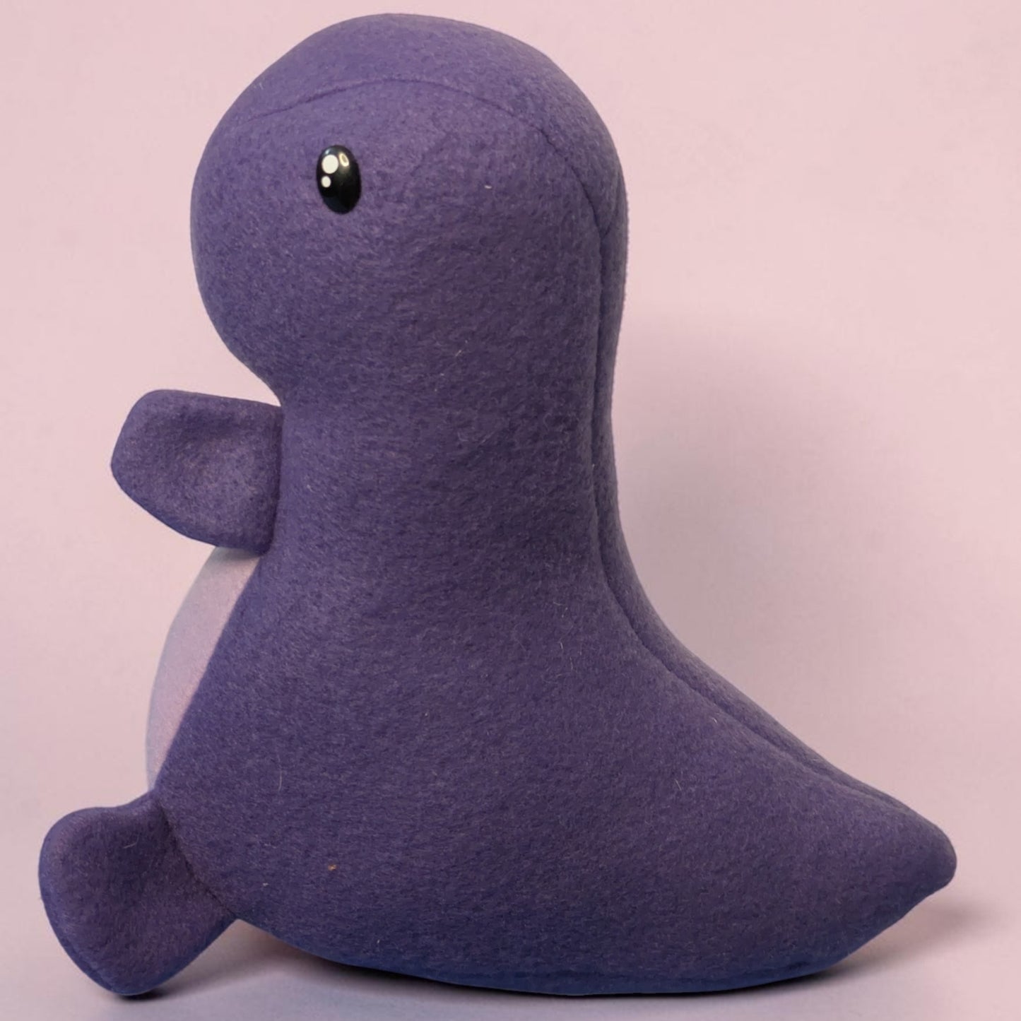 A small plush dinosaur toy made of fleece fabric in a muted purple color, with a glossy black eye and a pale underside, photographed against a plain light pink background.