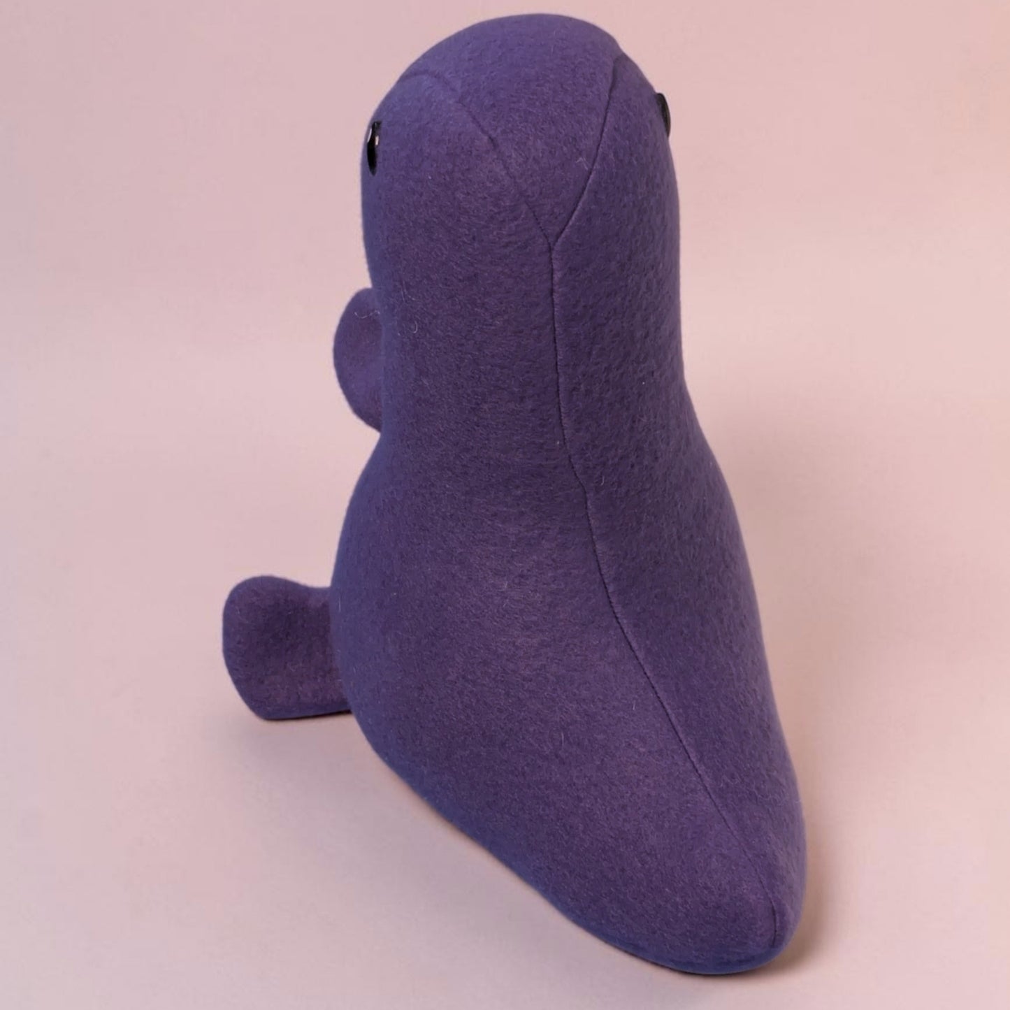 A purple fleece dinosaur plush toy with small black safety eyes, shown from the back angle on a solid pale pink background.