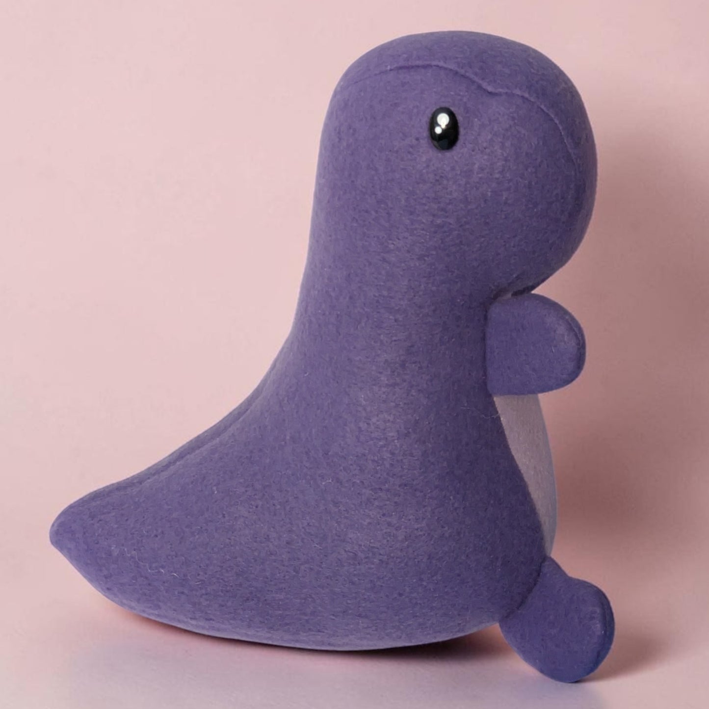 A purple fleece dinosaur plush toy with a glossy black safety eye, sitting against a solid light pink background.