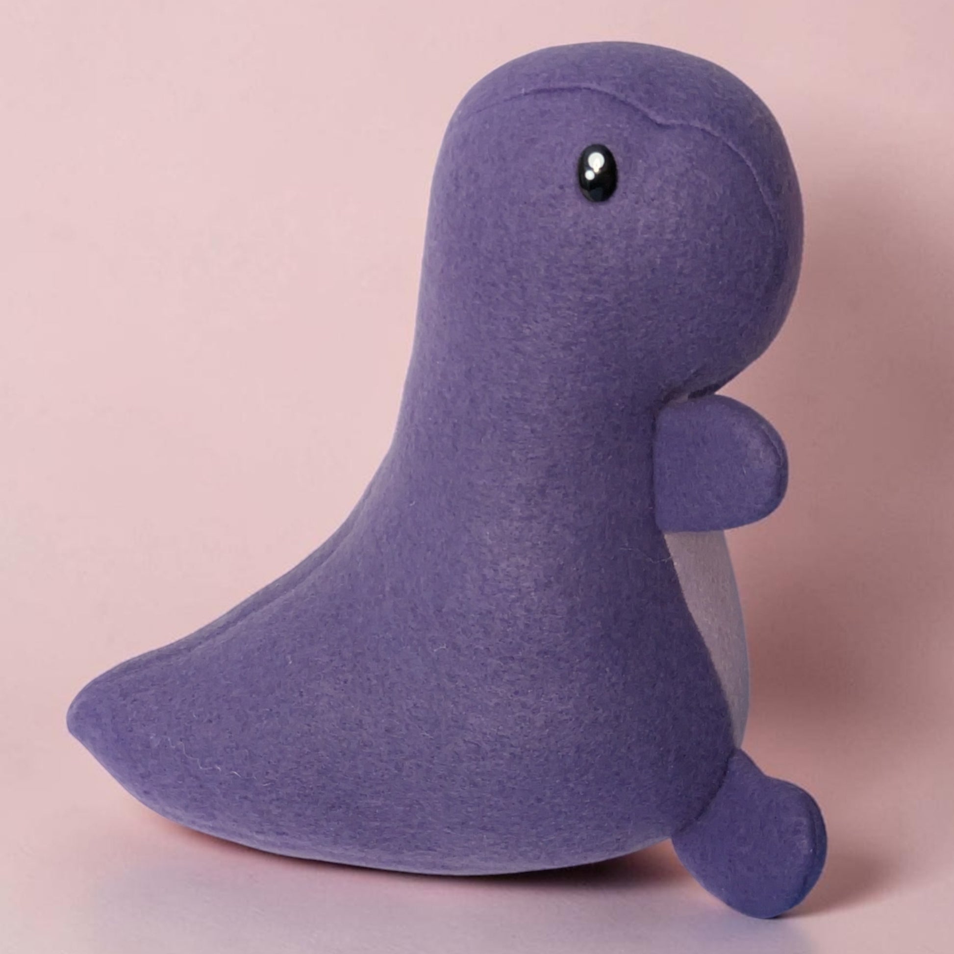 A purple fleece dinosaur plush toy with a glossy black safety eye, sitting against a solid light pink background.