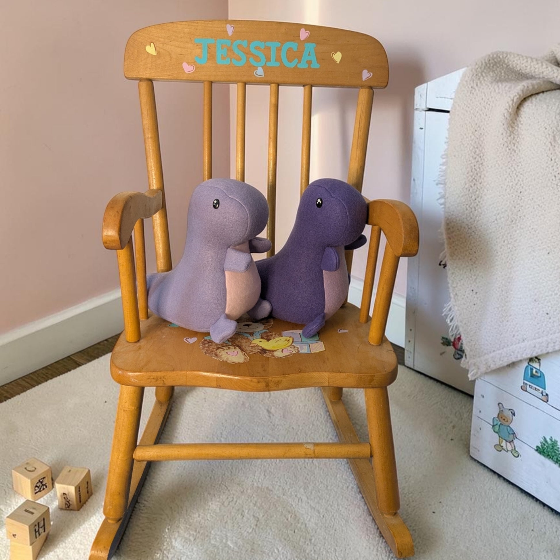 A wooden rocking chair in a child's room, personalized with the name 'JESSICA' on the backrest. Two plush dinosaur toys, one light purple and one dark purple, are sitting on the chair.