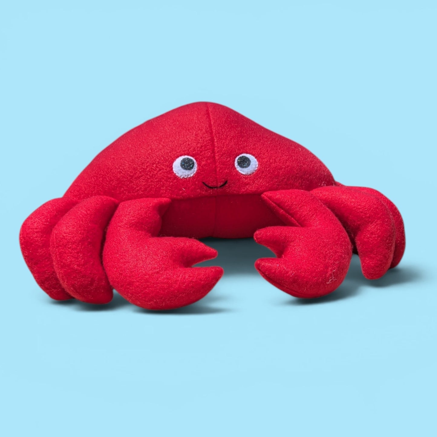 A red plush crab toy with felt eyes and a small smile, photographed on a light blue background.