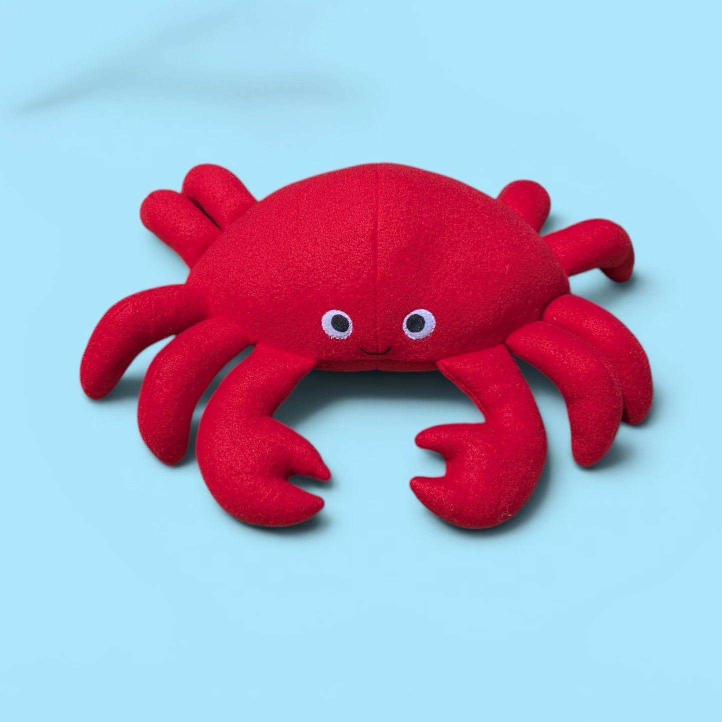 A red plush crab toy with felt eyes and a small smile, photographed on a light blue background.