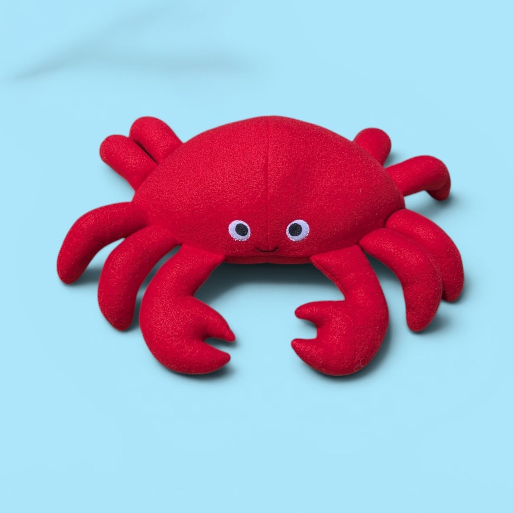 A red plush crab toy with felt eyes and a small smile, photographed on a light blue background.