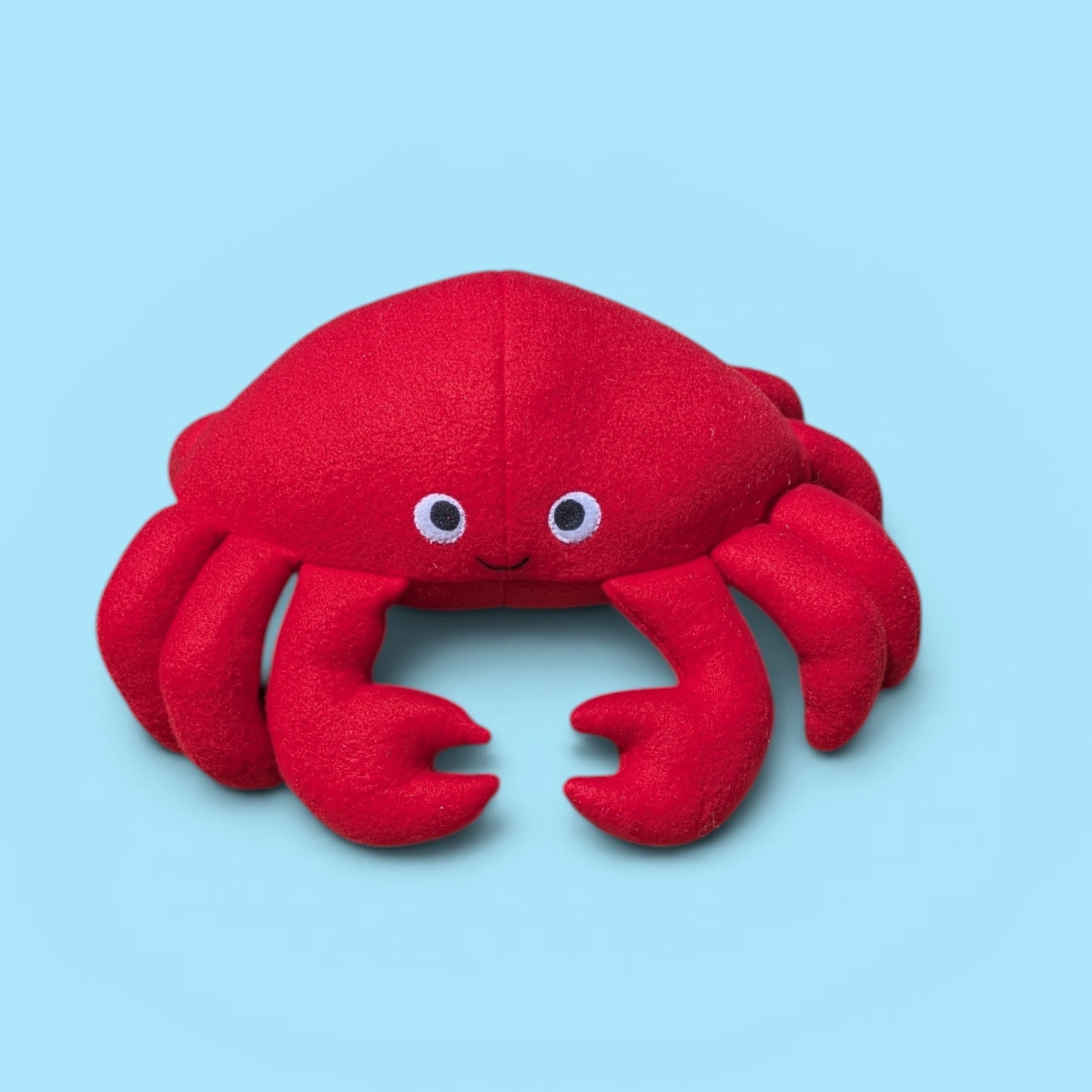 A red plush crab toy with felt eyes and a small smile, centered on a light blue background.