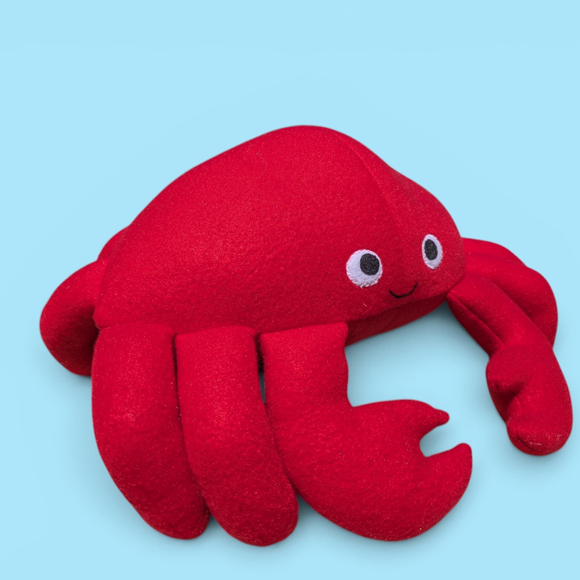 A red plush crab toy with stitched felt eyes and a smile, shown on a solid light blue background.