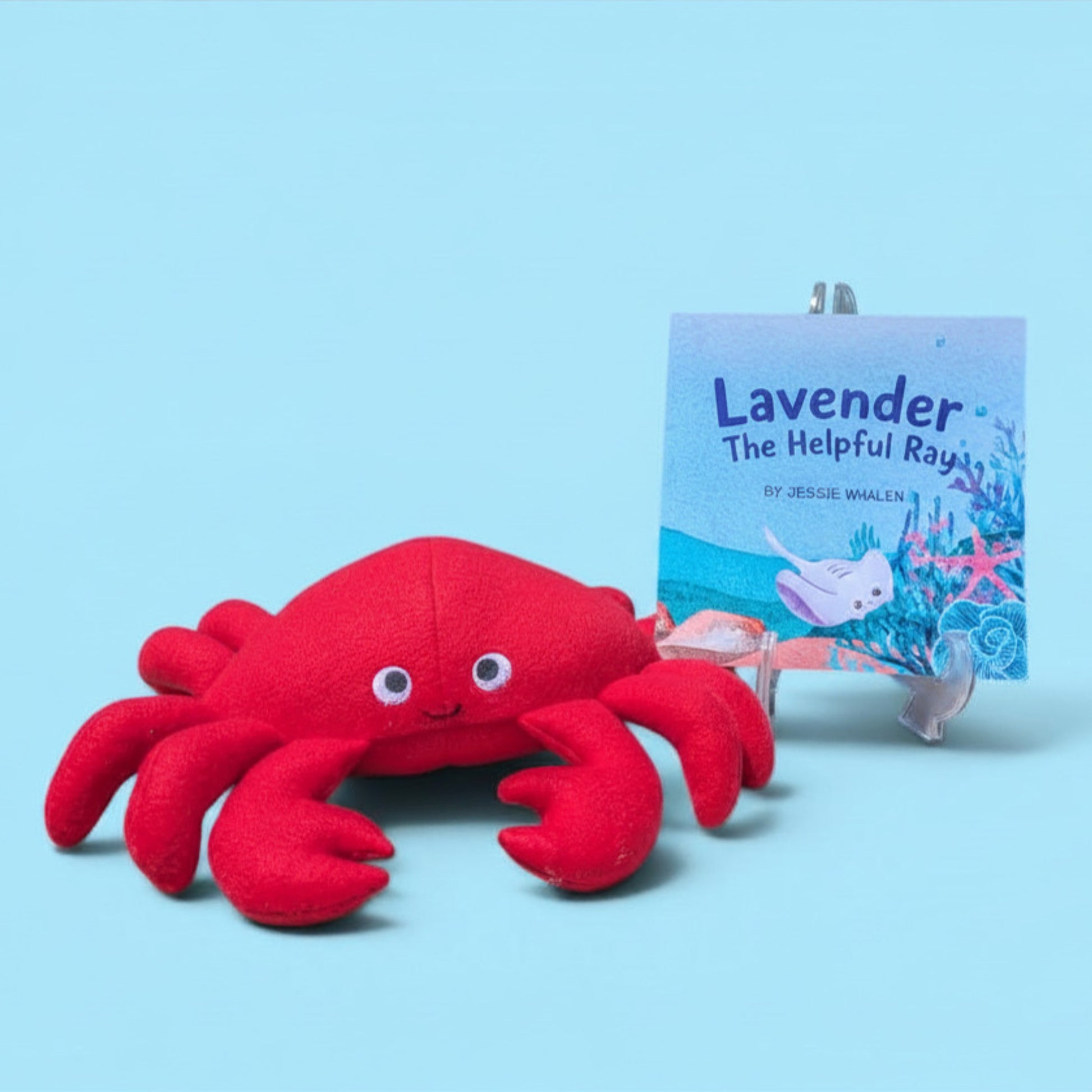 A red plush crab toy with felt eyes is shown in front of a children's book titled "Lavender The Helpful Ray" on a clear display stand, against a light blue background.