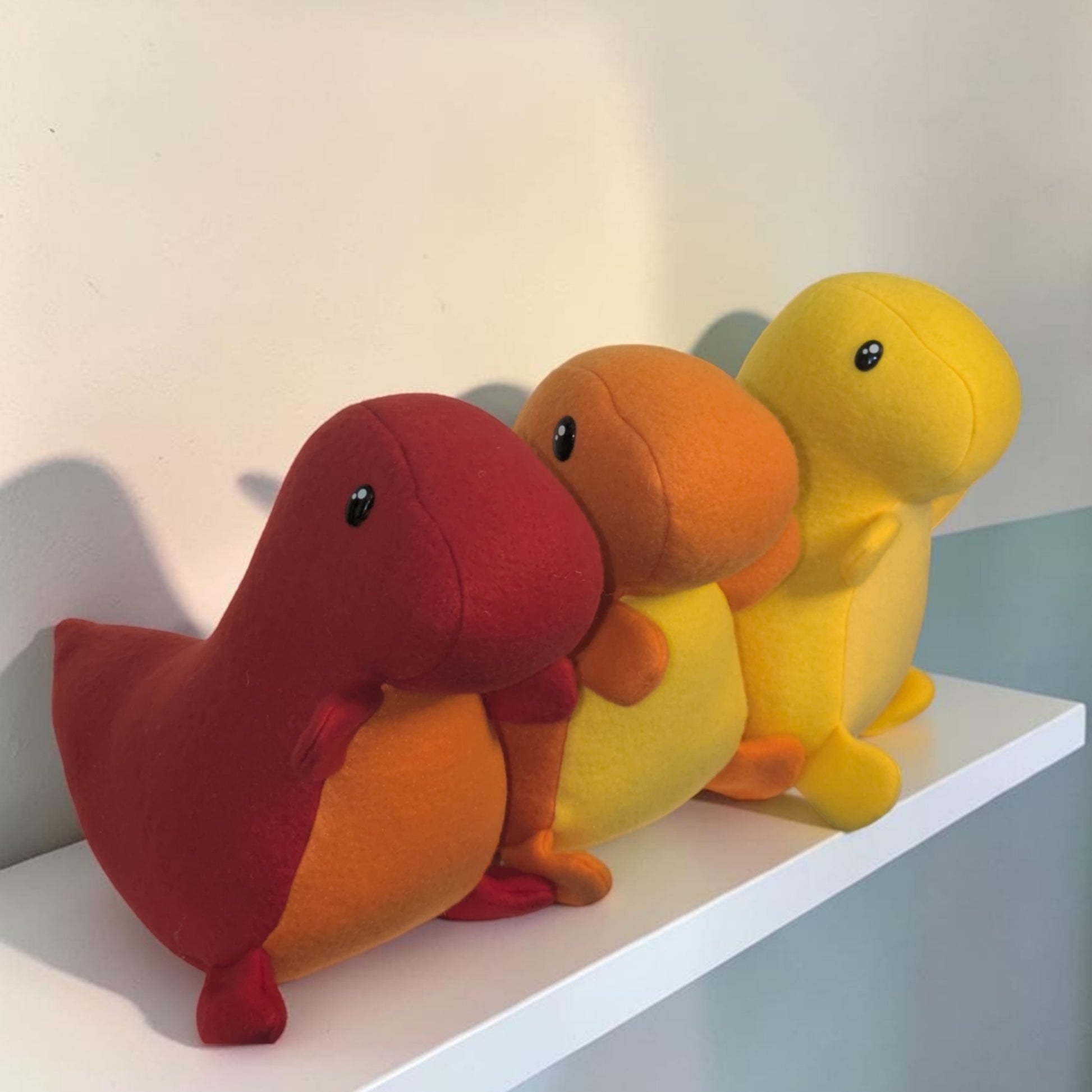 Three small plush dinosaur toys in red, orange, and yellow, arranged side by side on a white shelf against a plain wall.