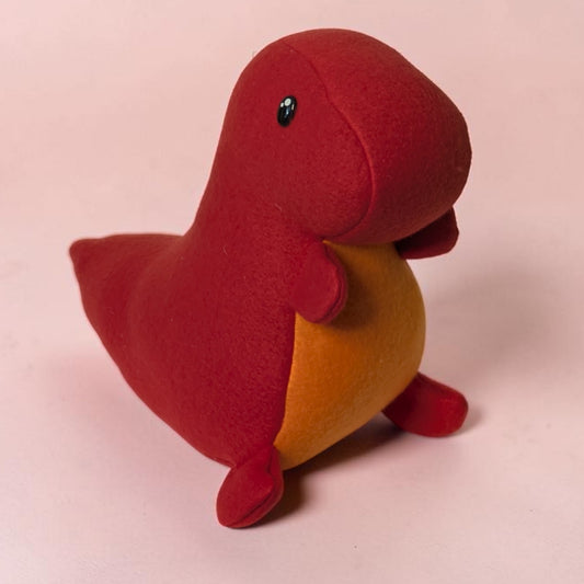 A small handmade plush dinosaur toy in red and orange, photographed on a plain light pink background.