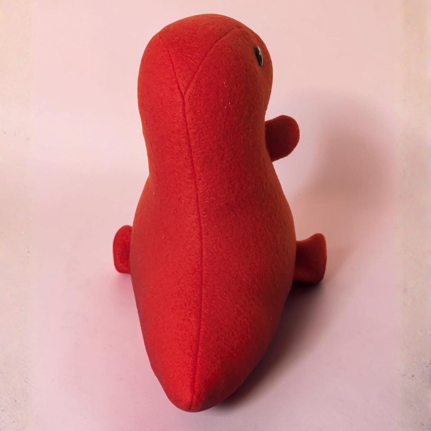 A red dinosaur plush toy shaped, photographed from the back  against a plain light pink background.