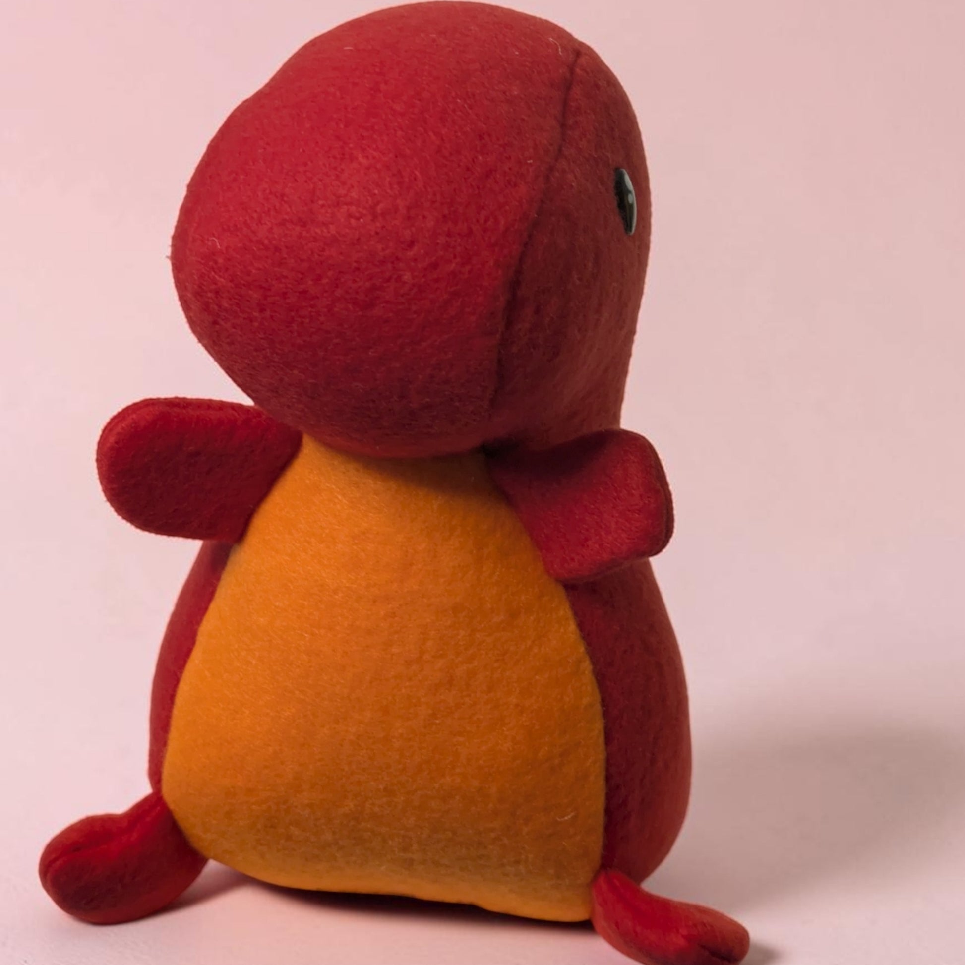 A small plush toy shaped like a rounded dinosaur. The stuffed animal has a dark red outer body with an orange belly panel and a tiny dark eye detail, photographed against a plain light pink background.