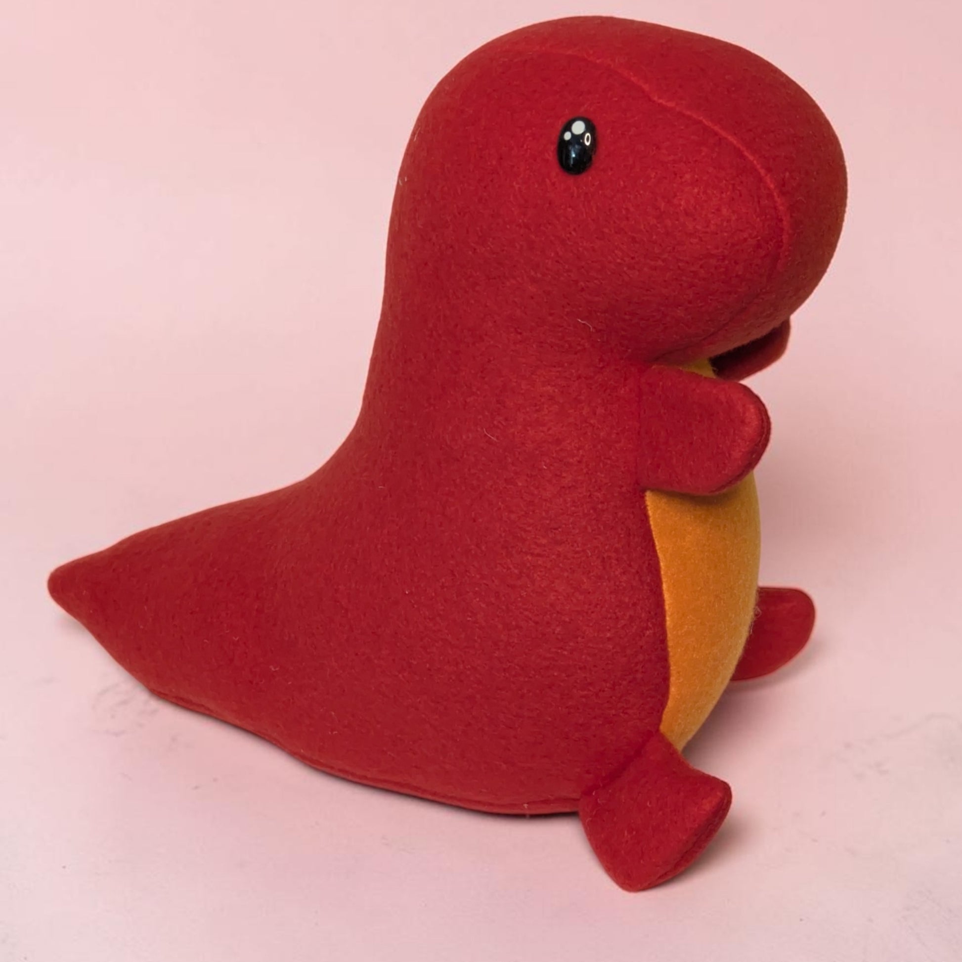 A red fleece dinosaur plush toy stuffed animal with a orange belly and small black eyes, sitting on a light pink background.