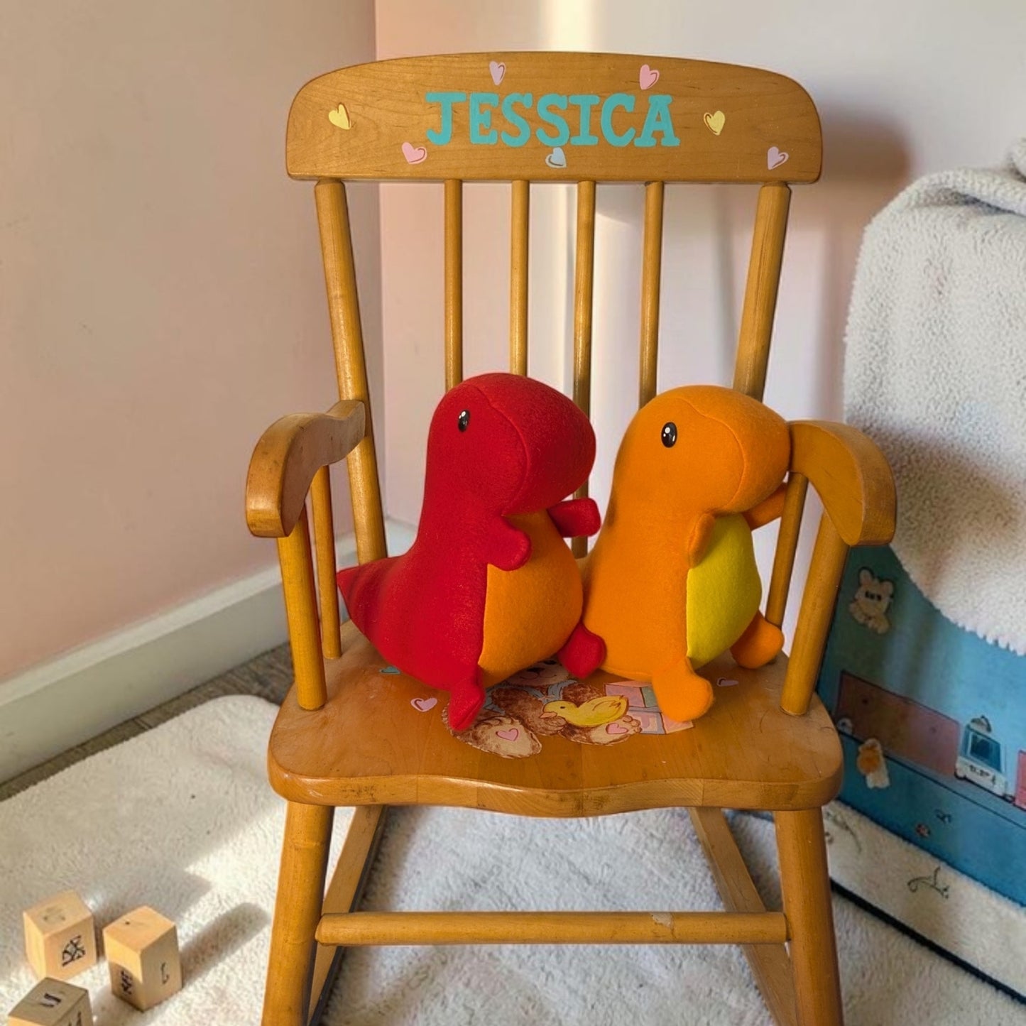 A wooden rocking chair in a child's room setting, featuring the name 'JESSICA' painted on the backrest. Two plush dinosaur toys, one red and one orange, are sitting on the chair.