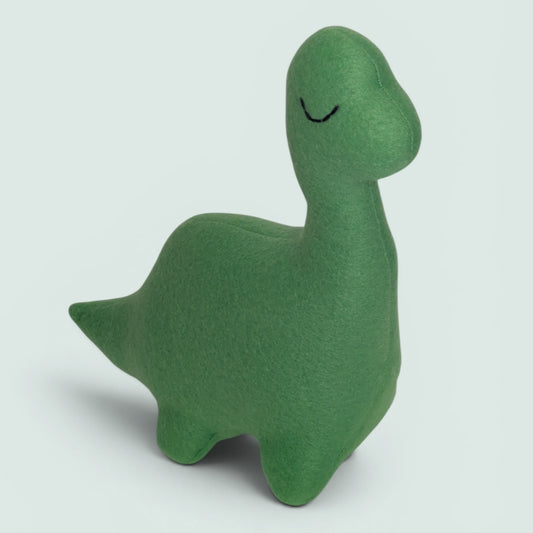 A small plush  dinosaur toy made of fleece fabric in green, with a small black embroidered eye, photographed against a solid pale blue background.