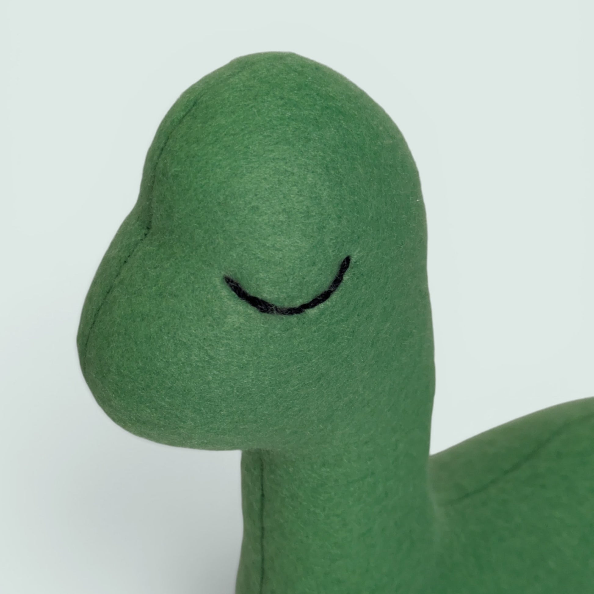 A cute green plush toy shaped like a small dinosaur featuring a soft fuzzy texture, a small black stitched eye, set against a solid pale blue background.