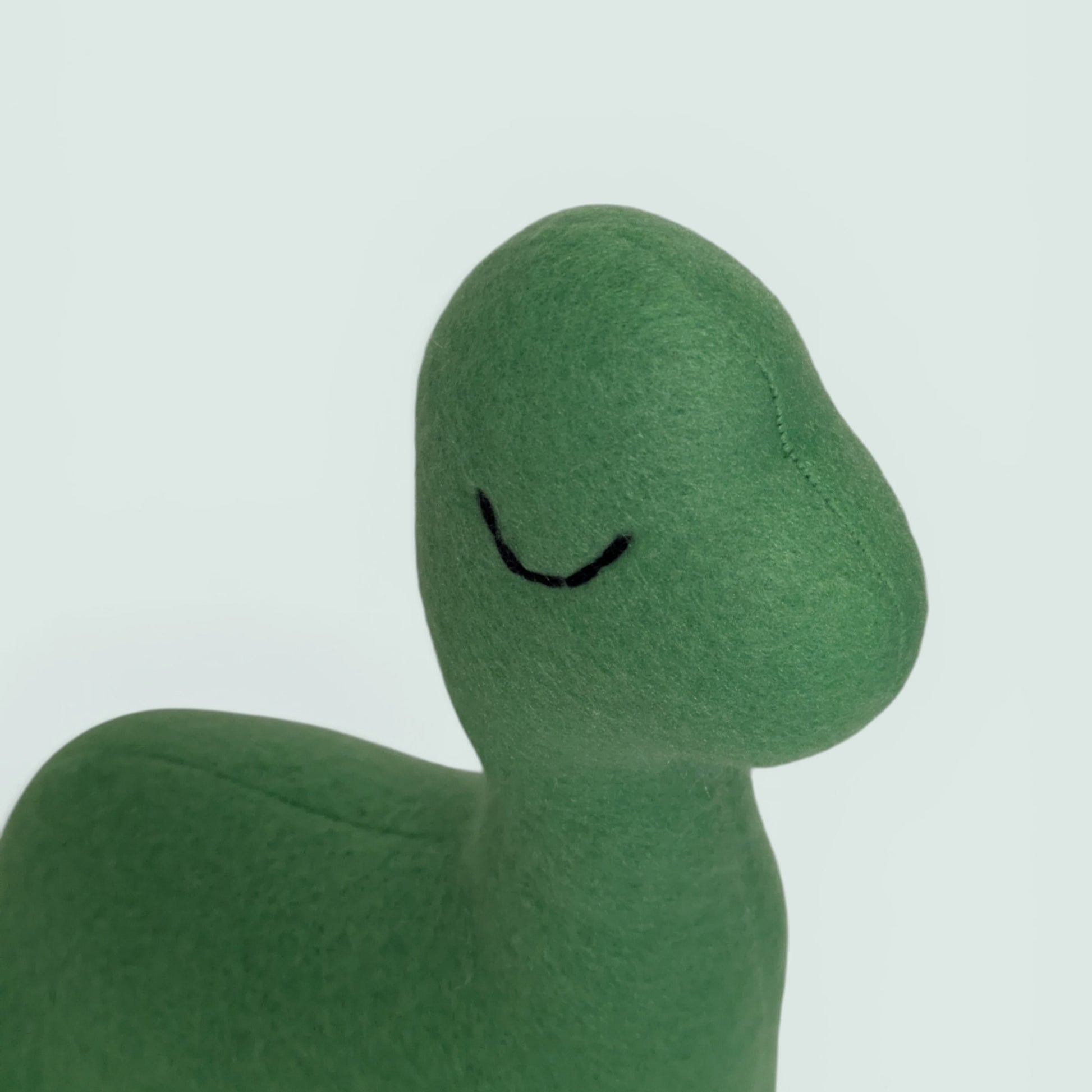 A close-up of a green fleece dinosaur toy with a simple black embroidered smiley face, set against a plain light gray background.