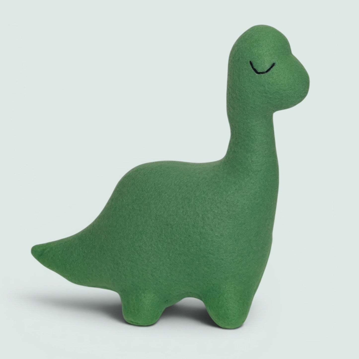 A green fleece dinosaur toy with a simple embroidered eye, standing against a light gray background.