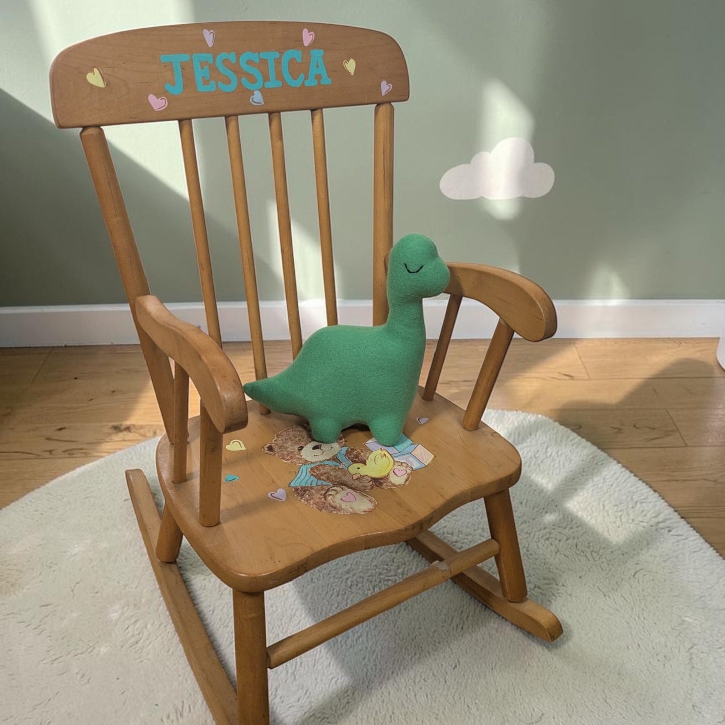 A wooden children's rocking chair with the name 'JESSICA' painted on the backrest, styled with a green plush dinosaur toy on the seat, set on a white rug in a child's room.