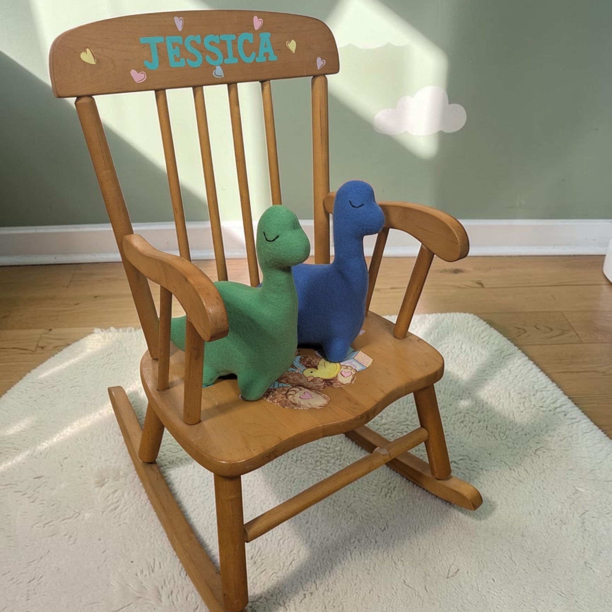 A wooden rocking chair with the name 'JESSICA' painted on the backrest, styled with two plush dinosaur toys on a white rug in a nursery setting.