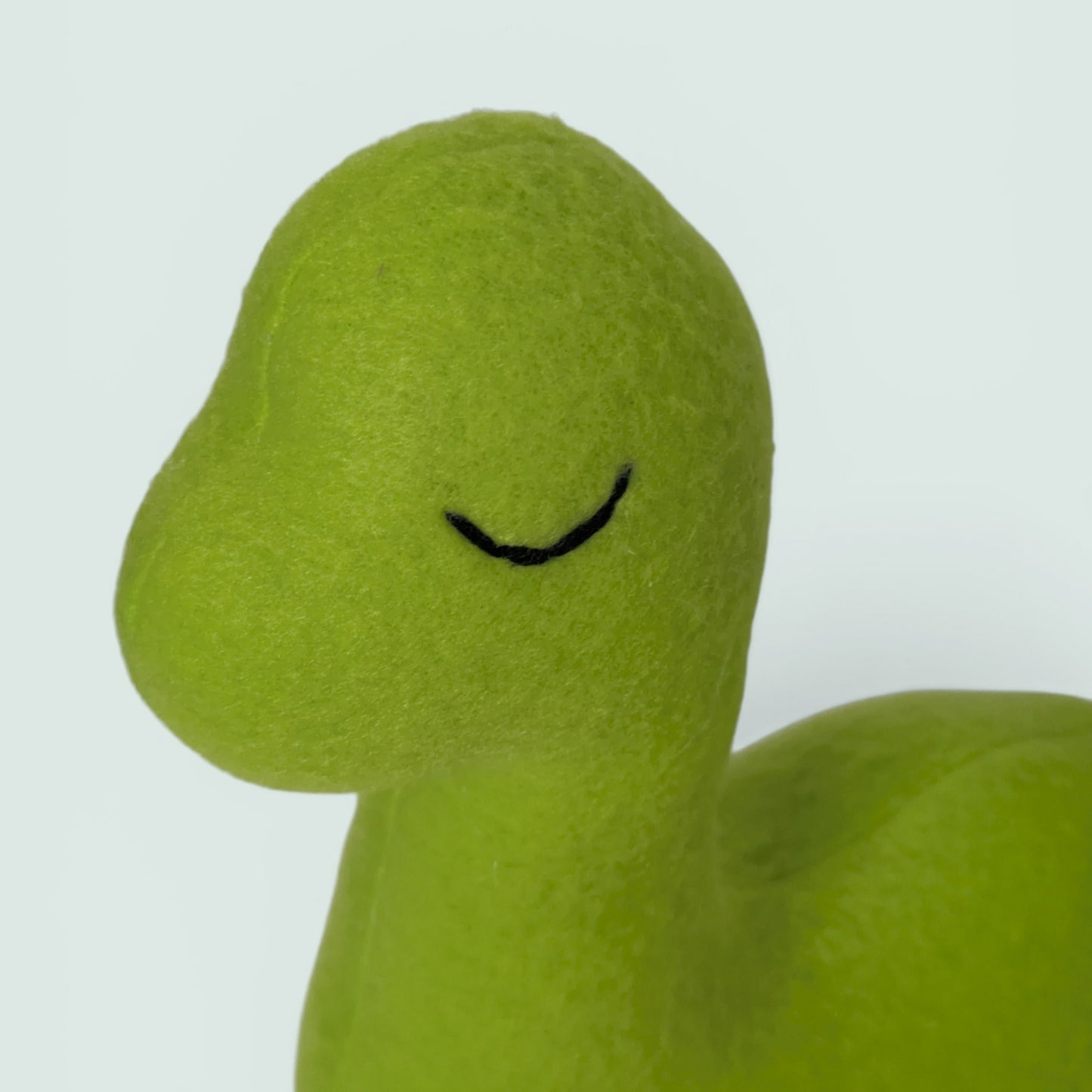 Close-up of a lime green fleece dinosaur toy with a simple black stitched eye on a light gray background.