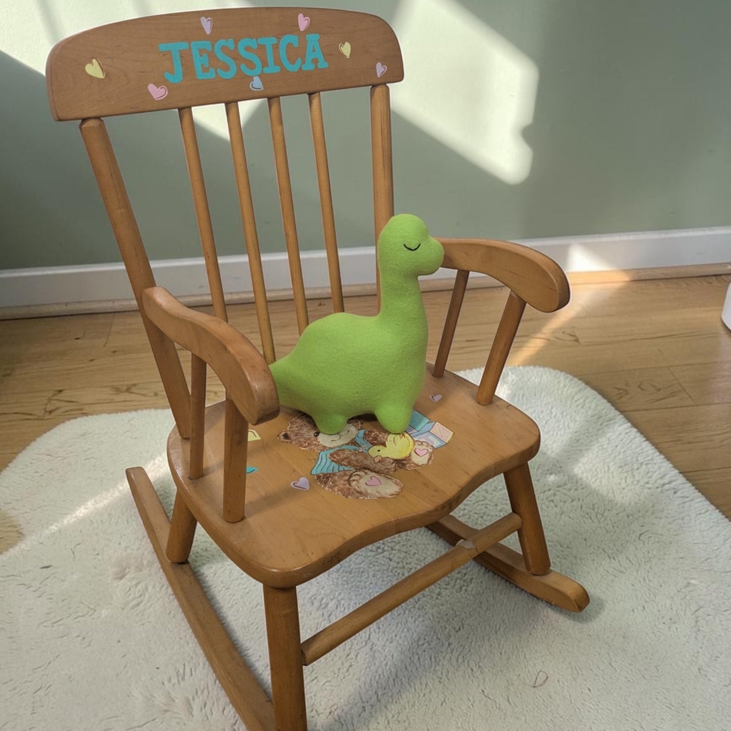 A wooden children's rocking chair with 'JESSICA' painted on the backrest, styled with a green dinosaur toy on the seat in a home setting.