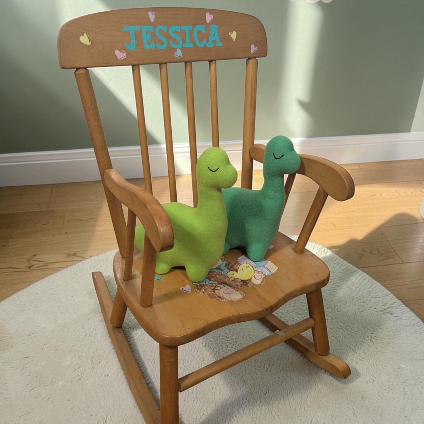 A wooden rocking chair with the name 'JESSICA' and small heart details on the backrest, styled with two plush dinosaur toys on a white rug in a room with a green wall and wood floor.