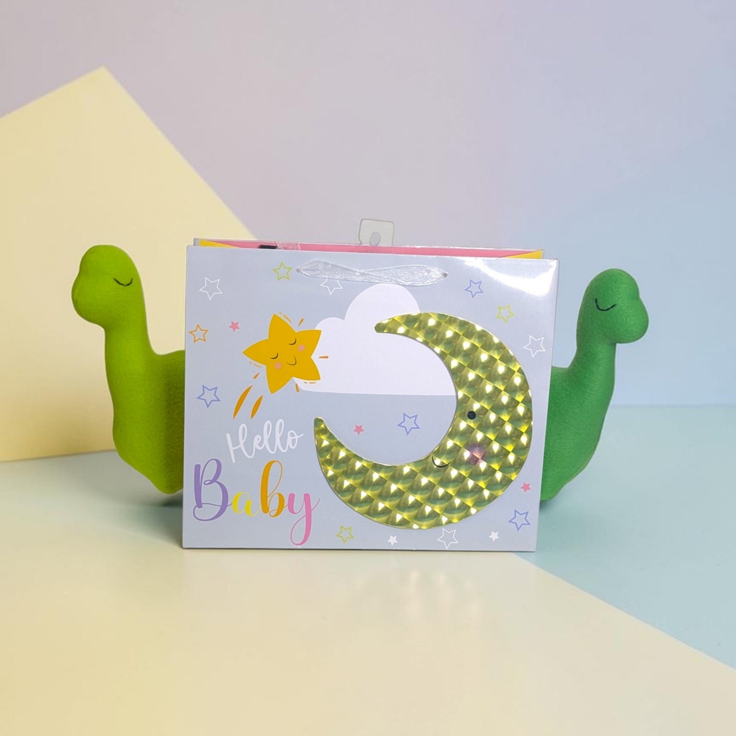 A baby shower gift box with a large glittery gold crescent moon and 'Hello Baby' text, flanked by two green dinosaur figures on a pastel-colored background.