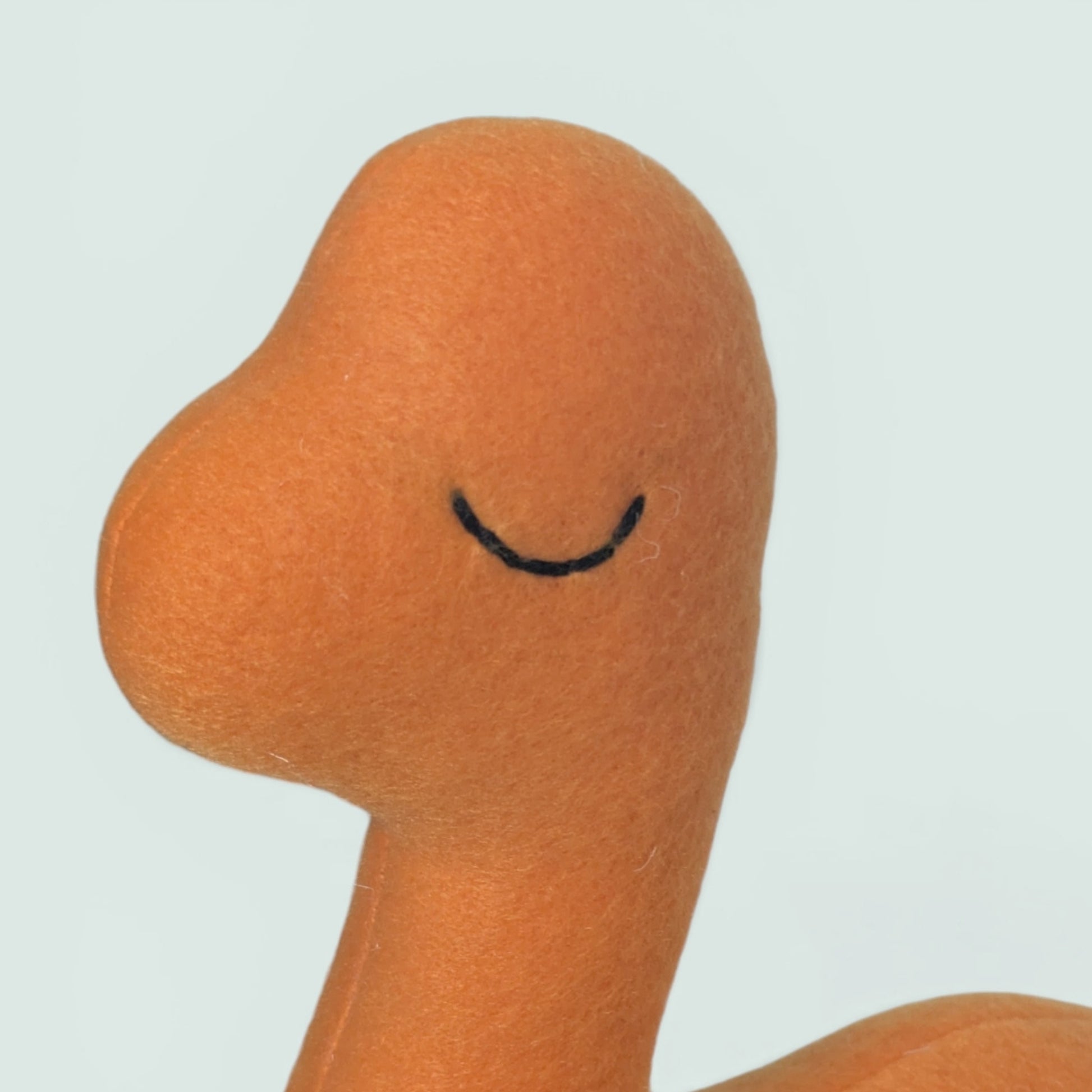 Close-up of a soft orange plush toy head with a simple black stitched eye on a pale blue background.