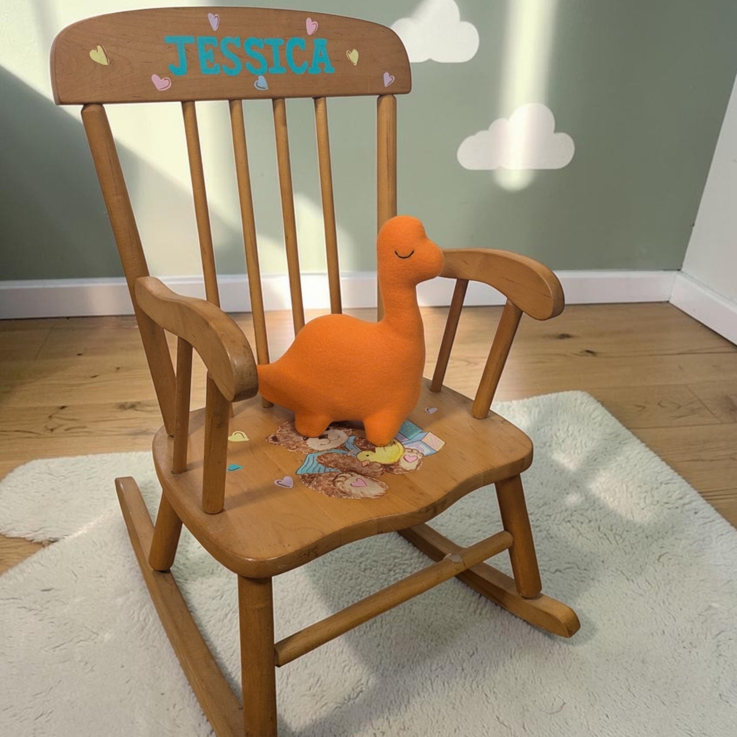 A wooden children's rocking chair in a nursery setting, featuring a personalized backrest with the name 'JESSICA' in blue letters and small heart decals. An orange plush dinosaur toy sits on the chair's seat, which is decorated with small stickers.