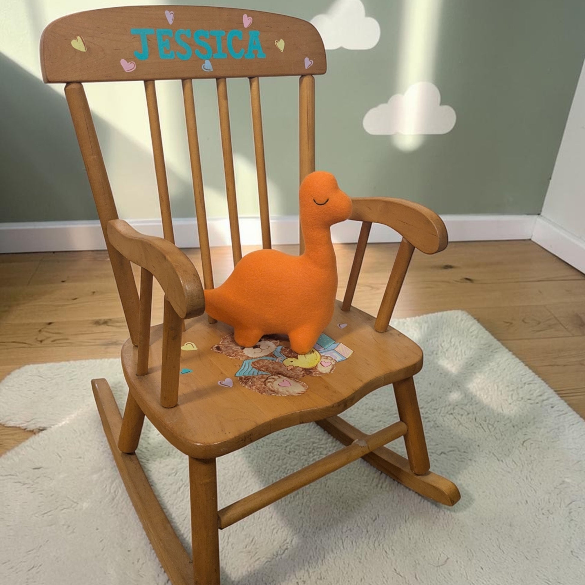 A wooden children's rocking chair in a nursery setting, featuring a personalized backrest with the name 'JESSICA' in blue letters and small heart decals. An orange plush dinosaur toy sits on the chair's seat, which is decorated with small stickers.