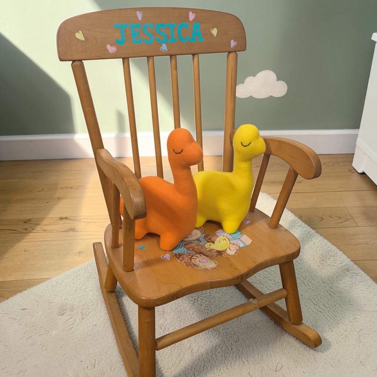 A wooden children's rocking chair with the name 'JESSICA' painted on the backrest, styled with two plush dinosaur toys on a white rug in a child's room.