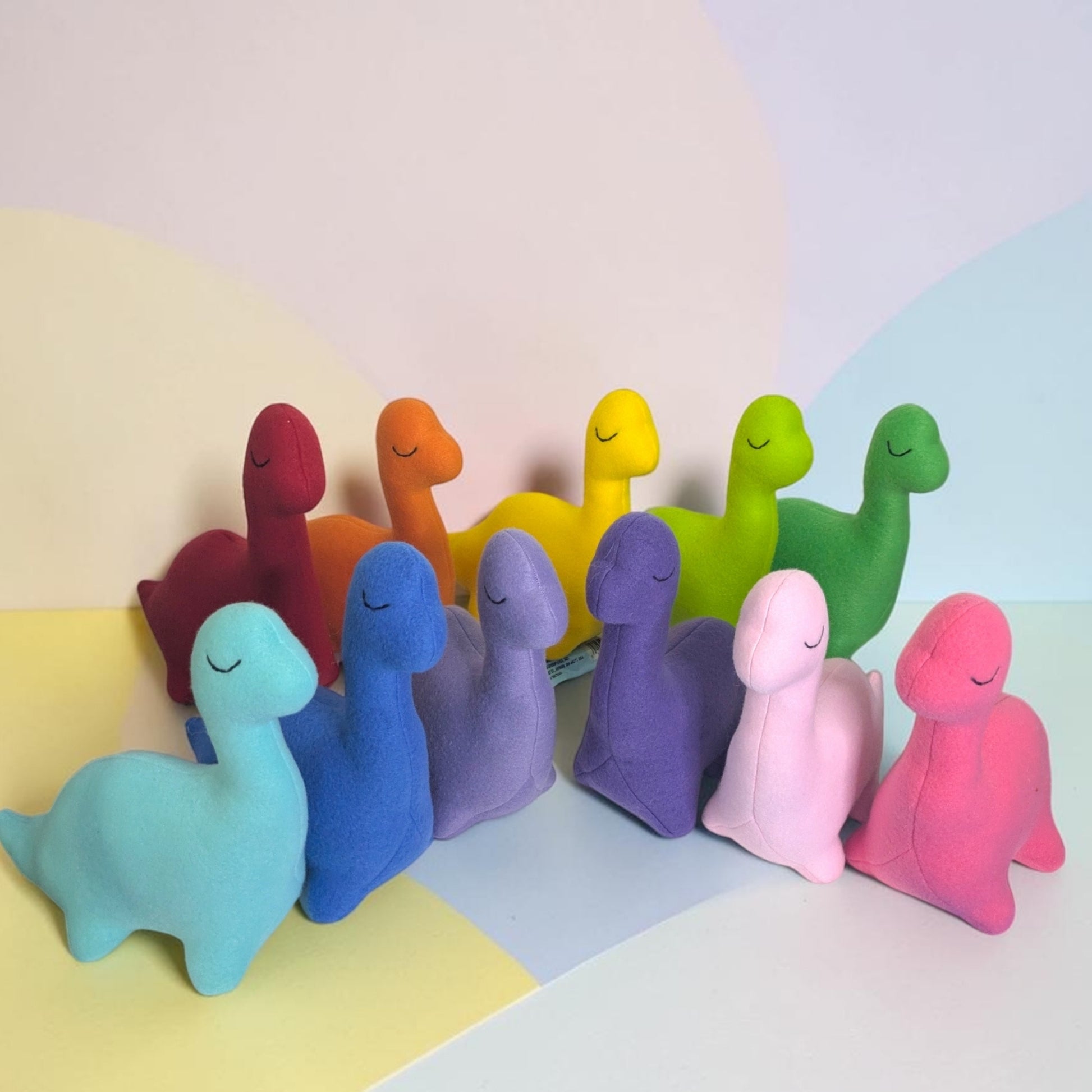 A group of small plush dinosaur toys in multiple pastel and bright colors, including light blue, dark blue, lavender, purple, pink, red, orange, yellow, green, and light green, arranged on a pastel-colored surface with a matching abstract background.