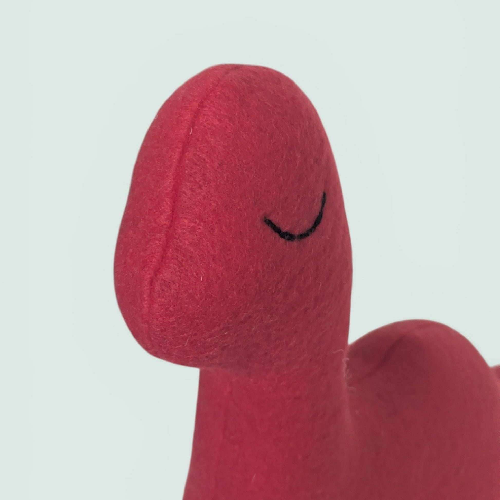Close-up of a red fleece dinosaur toy with a simple black embroidered eye, set against a light gray background.