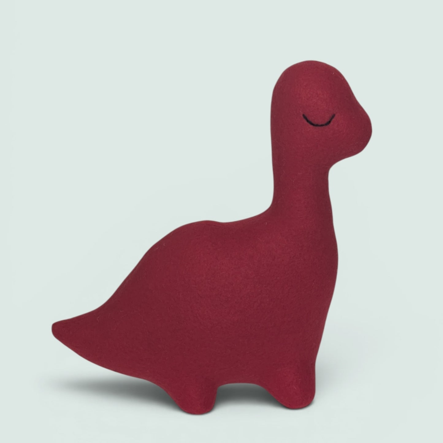 A plush dinosaur toy in a deep burgundy red color, featuring a simple embroidered closed-eye design on its head, set against a solid pale gray background.