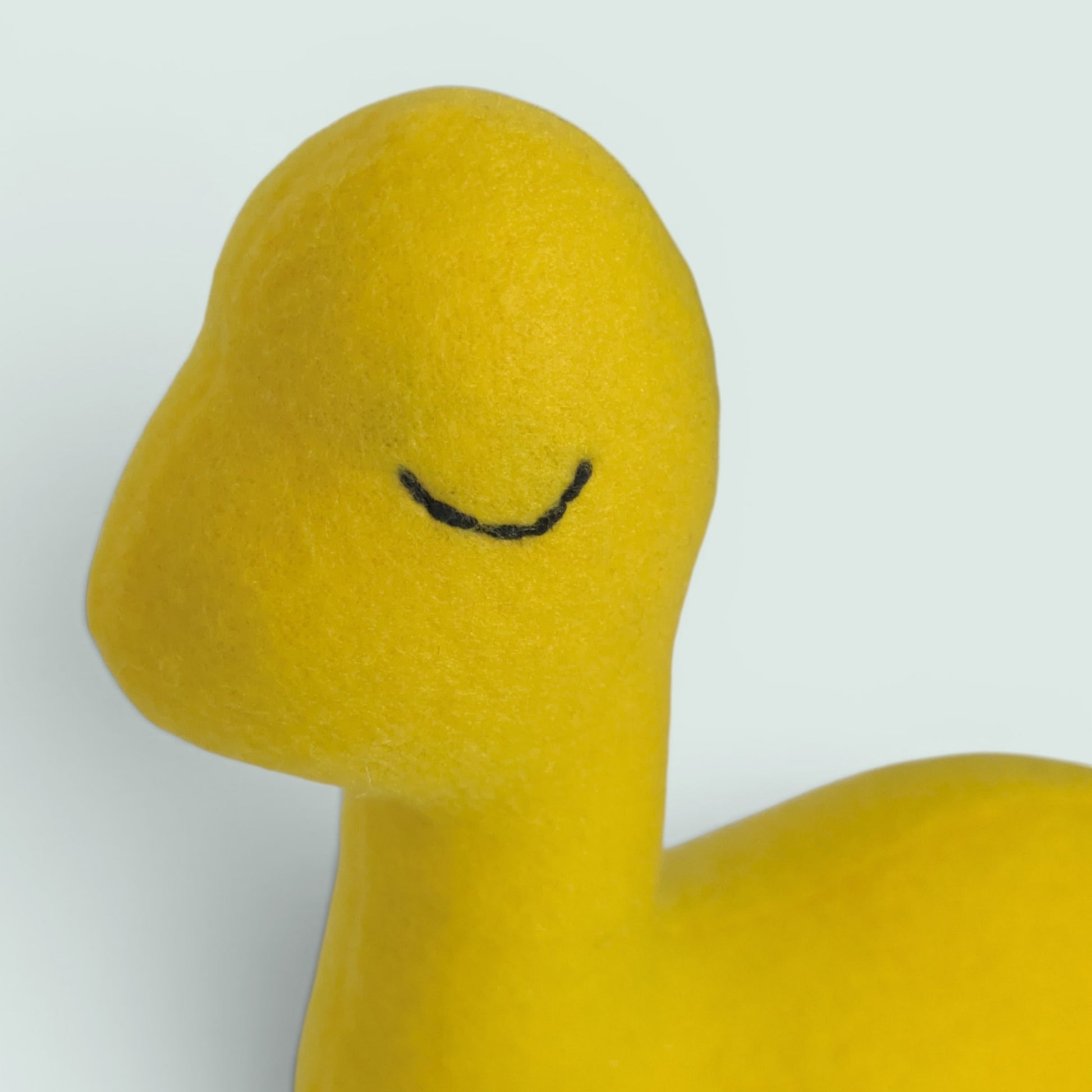 Close-up of a bright yellow dinosaur-shaped plush with a simple black curved line for a closed eye, shown against a plain light background.