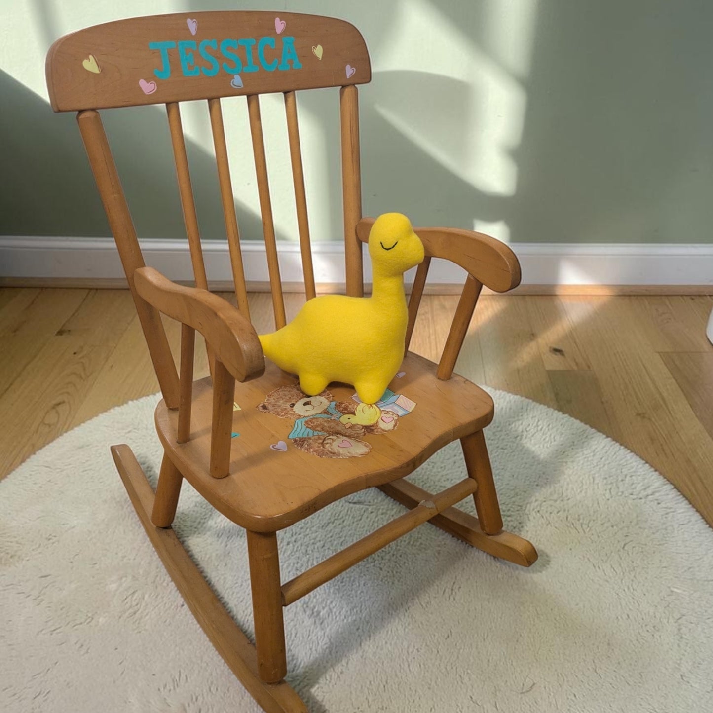 A wooden rocking chair with 'JESSICA' and heart details on the backrest, styled with a yellow plush dinosaur toy on the seat, set on a white round rug in a room with light wood floors and a green wall.
