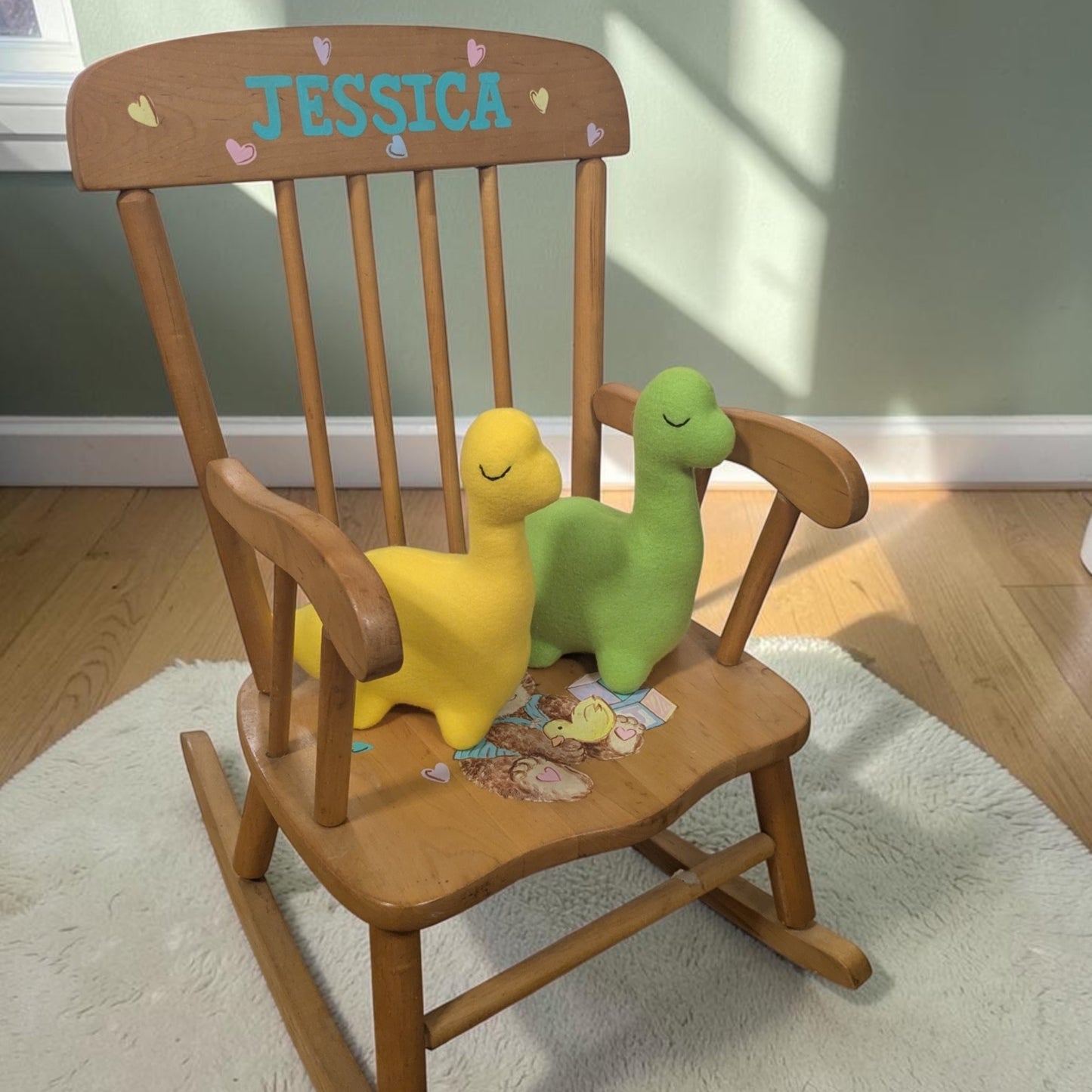 A wooden children's rocking chair with the name 'JESSICA' painted in blue on the backrest, styled with two plush dinosaur toys, one yellow and one green, on a white rug in a nursery setting.