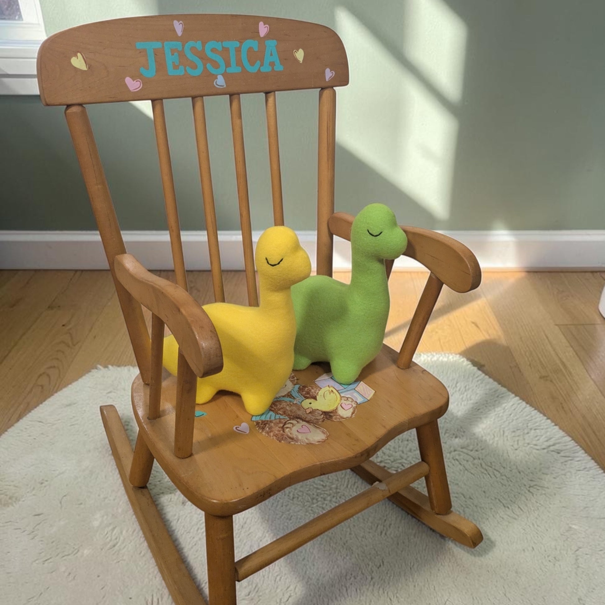 A wooden children's rocking chair with the name 'JESSICA' painted in blue on the backrest, styled with two plush dinosaur toys, one yellow and one green, on a white rug in a nursery setting.