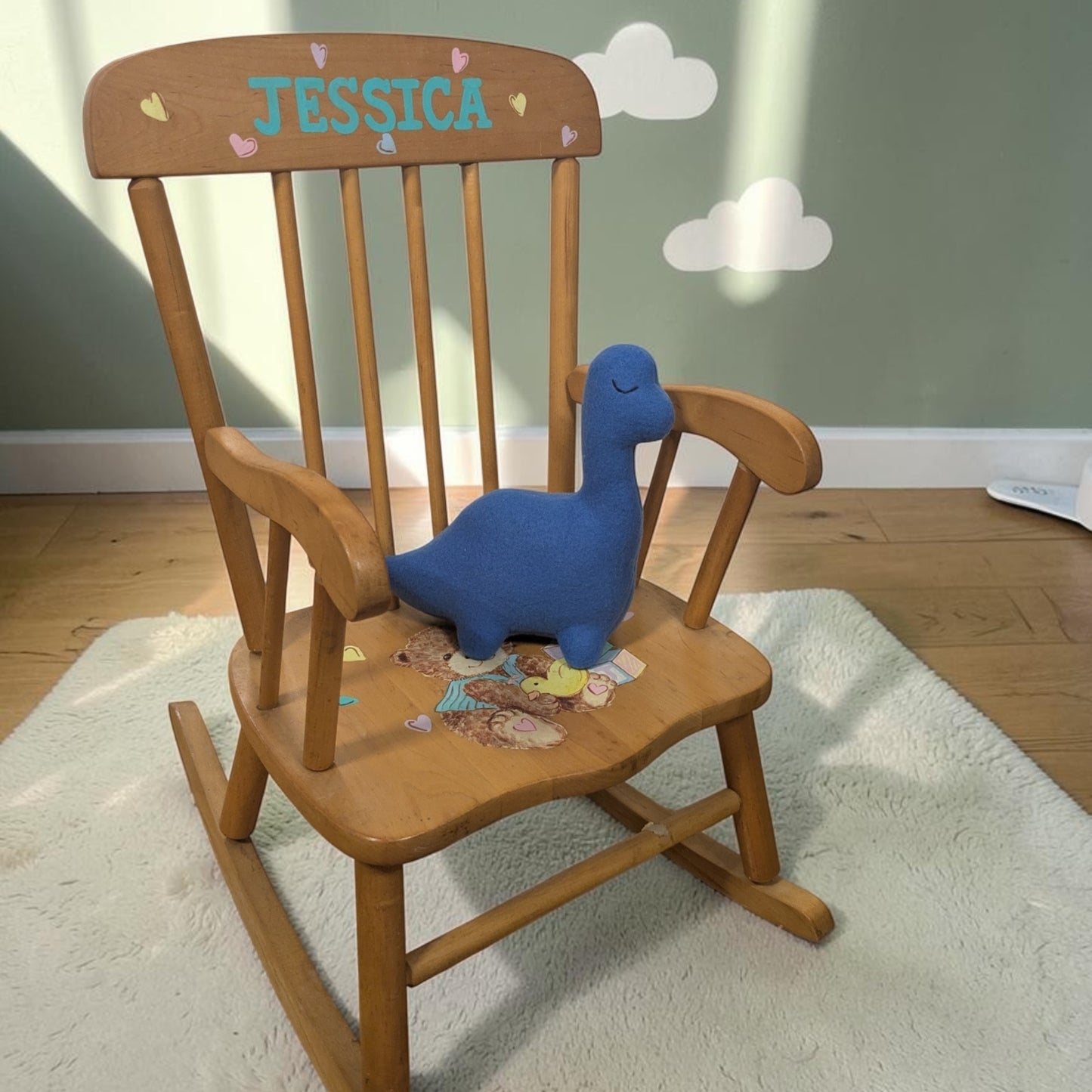 A wooden children's rocking chair with 'JESSICA' personalized on the backrest, featuring a blue plush dinosaur toy on the seat, set on a white rug in a child's room.