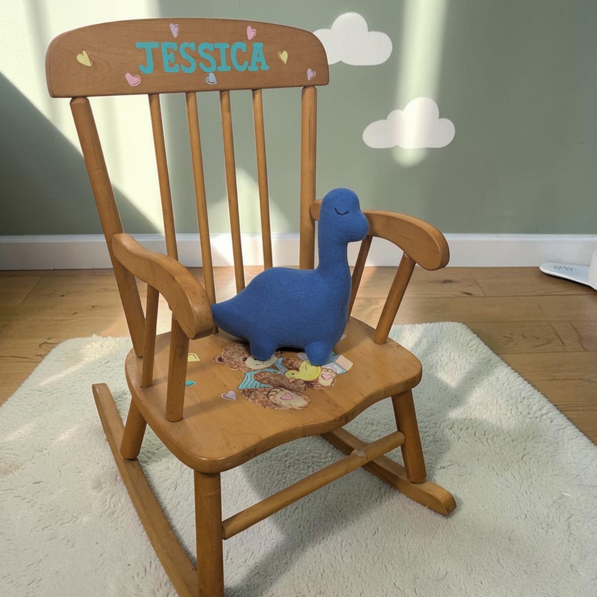 A wooden children's rocking chair with 'JESSICA' personalized on the backrest, featuring a blue plush dinosaur toy on the seat, set on a white rug in a child's room.