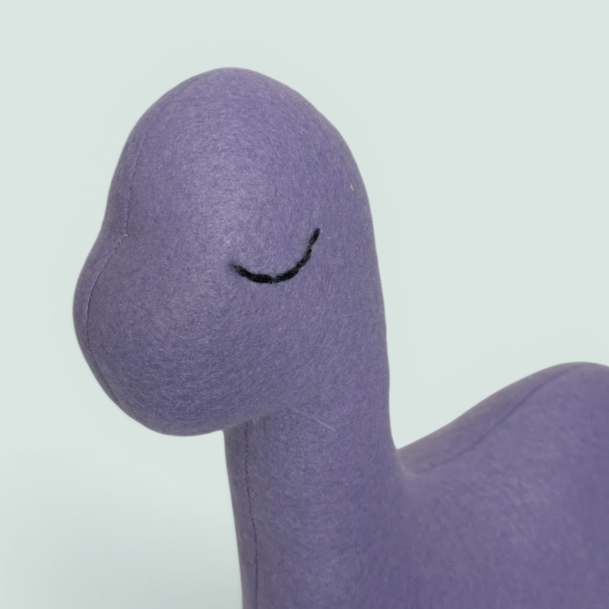 A close-up of a purple fleece plush toy with a simple black embroidered sleeping eye, set against a plain light gray background.
