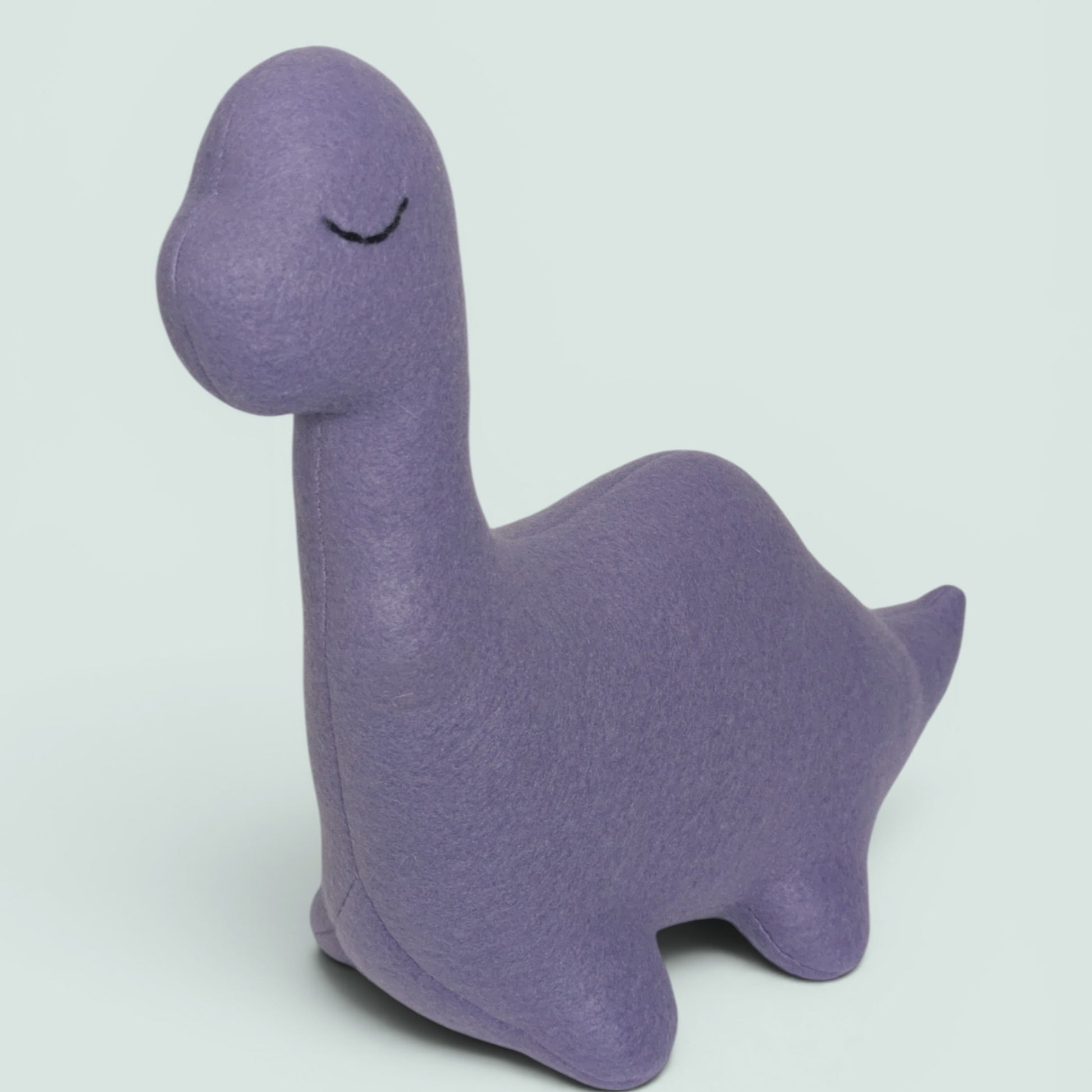 A lavender plush toy shaped like a stylized dinosaur, photographed from the side on a plain light background.
