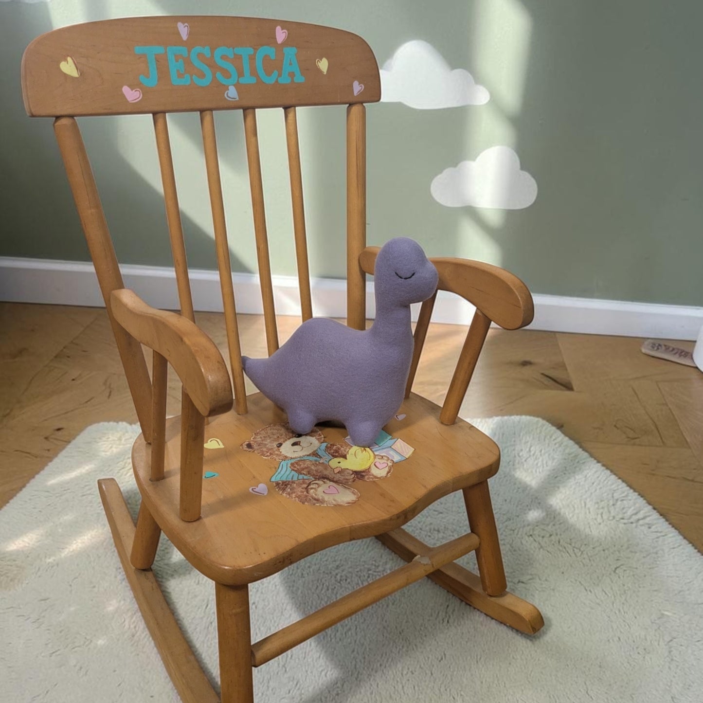 A wooden rocking chair with the name 'JESSICA' painted on the backrest and a purple plush dinosaur toy placed on the seat, set in a child's room on a white rug.