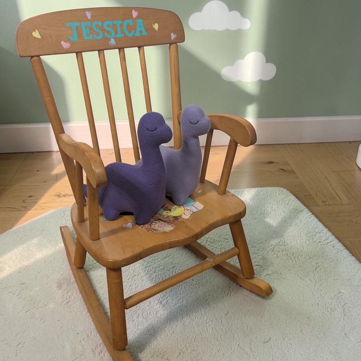 A wooden rocking chair with the name 'JESSICA' and small heart details on the backrest, styled with two purple plush dinosaur toys on a white rug in a child's room.